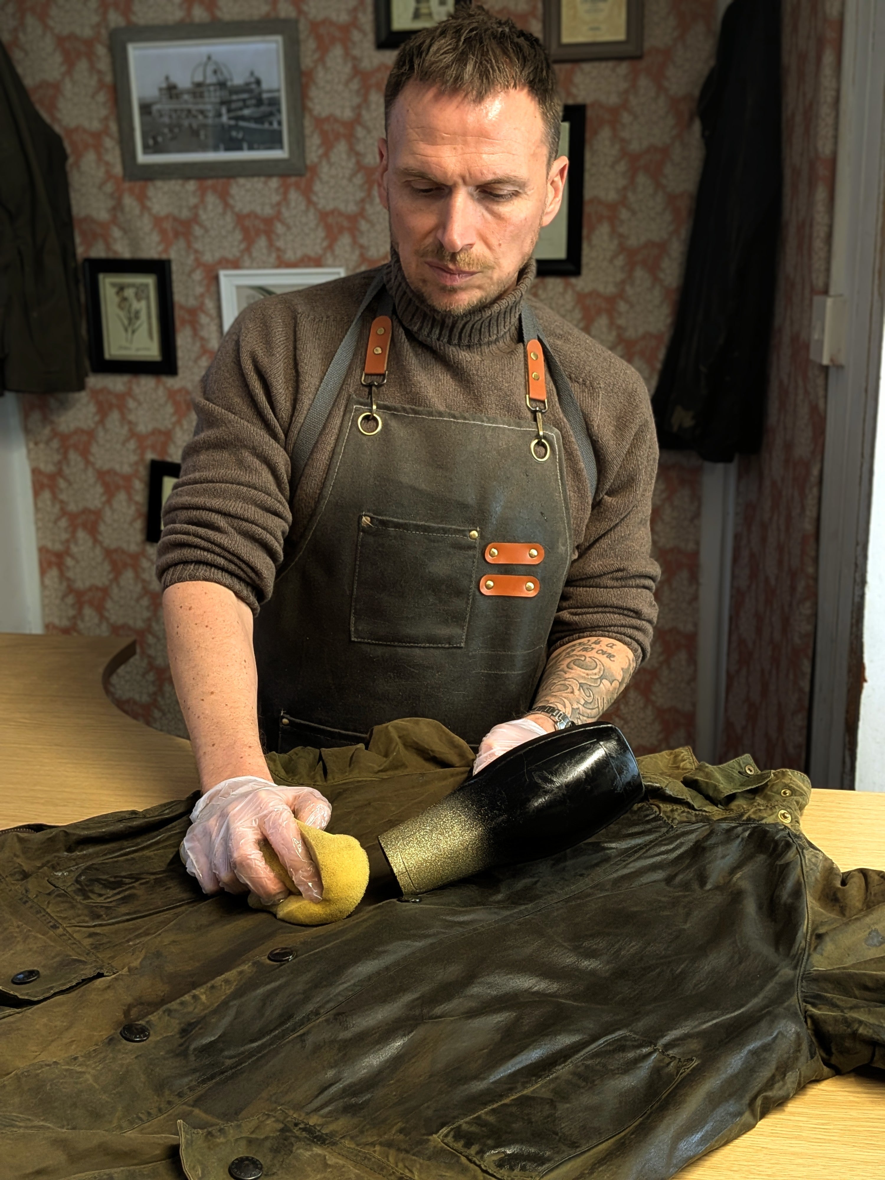 A man wearing an apron and gloves uses a sponge and a hair dryer to treat or restore a brown leather jacket on a table in a workshop. Framed photos and patterned wallpaper are visible in the background.