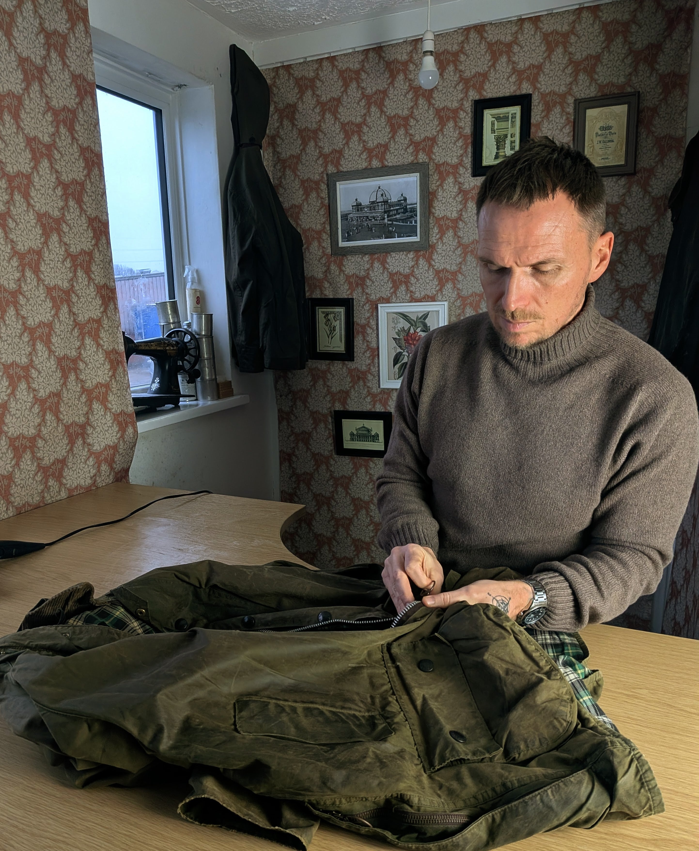 A man in a gray sweater sits at a wooden table, focused on repairing a green jacket. The room has patterned wallpaper, framed pictures on the wall, and a window letting in daylight.