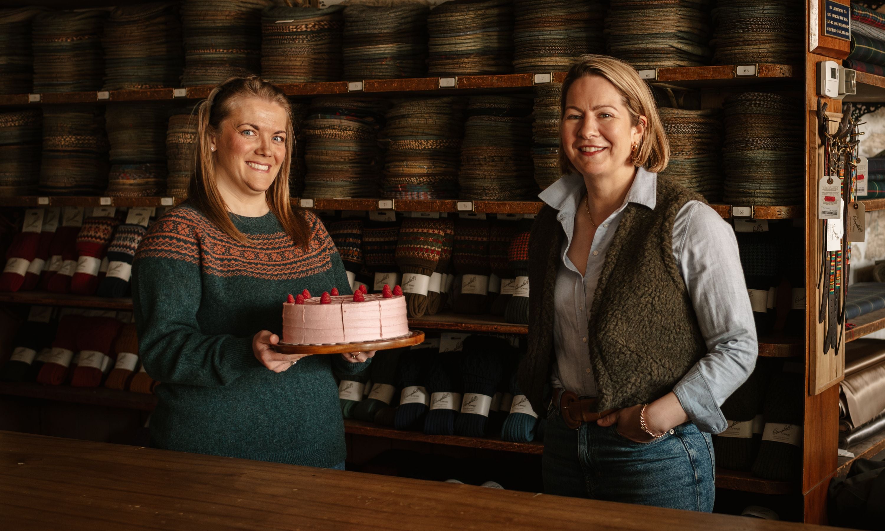 Two women stand in a yarn shop; one holds a pink frosted cake with raspberries. Shelves full of colorful yarn skeins line the wall behind them. Both women are smiling at the camera.