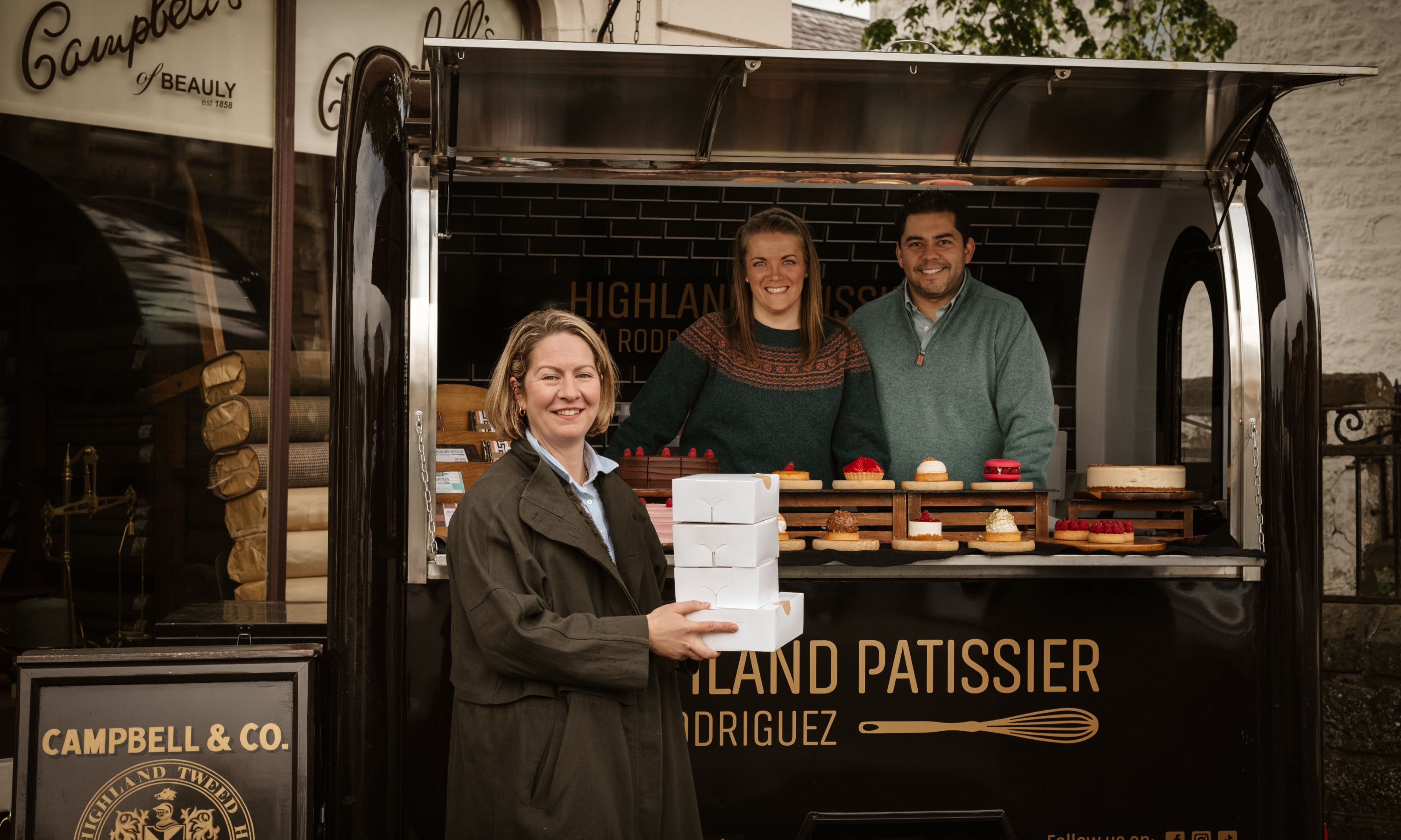 A smiling woman holding stacked pastry boxes stands in front of a food truck labeled “Highland Patissier.” Two people inside the truck smile behind a display of pastries and baked goods.