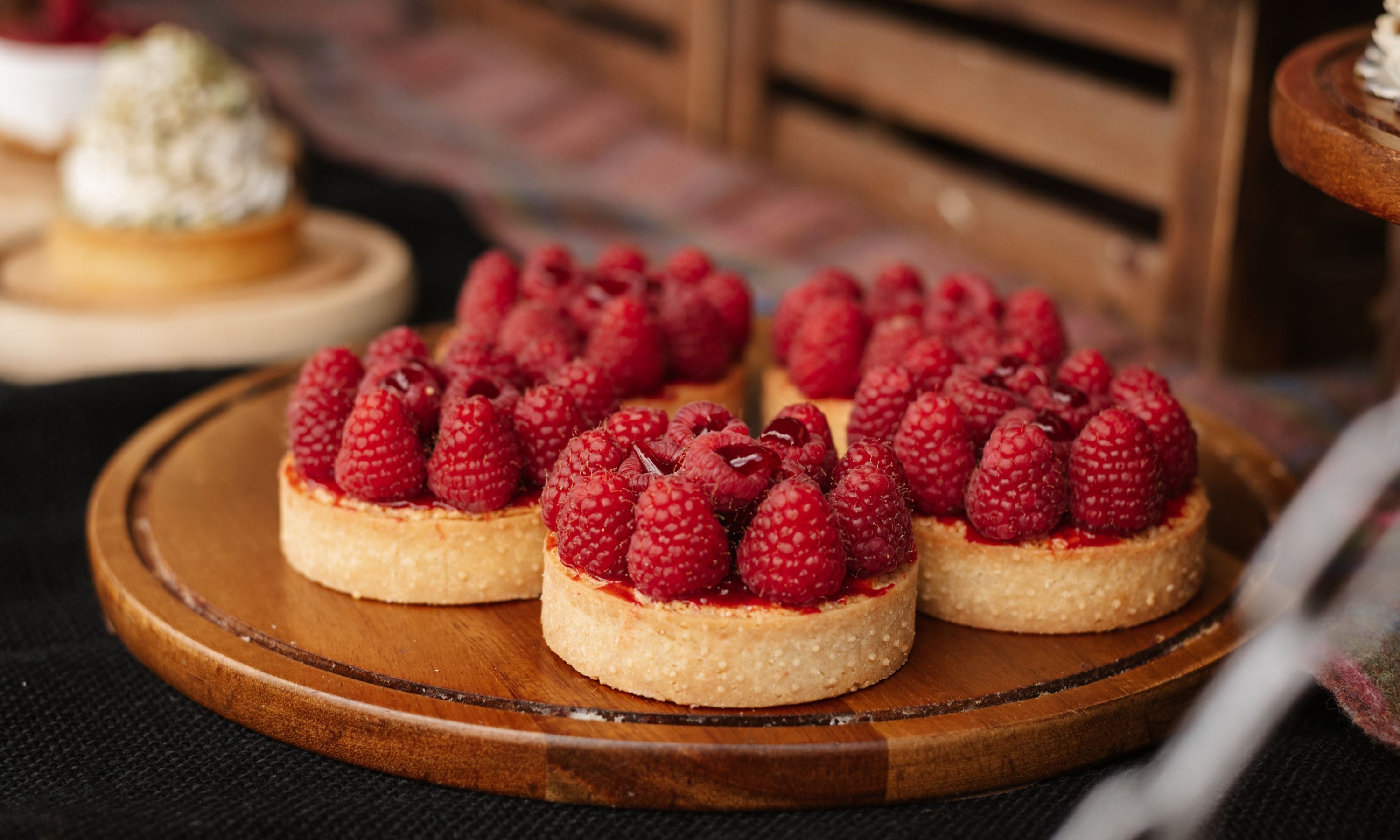 Three raspberry tarts with golden crusts and fresh raspberries on top are arranged on a wooden serving board. The background is softly blurred, suggesting a cozy, rustic setting.