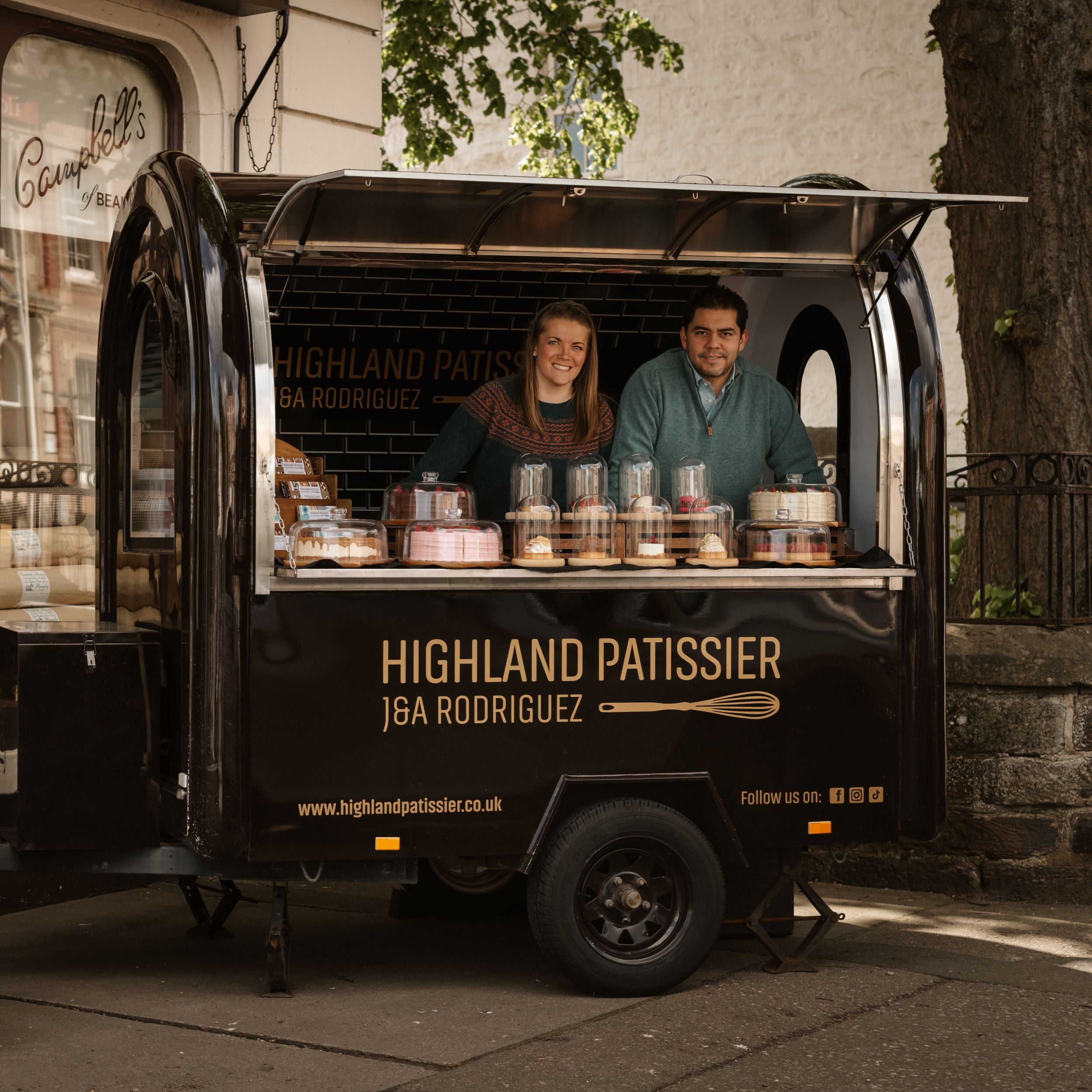 Two people smile from inside a black food trailer labeled Highland Patissier J&A Rodriguez, displaying various cakes and pastries on the counter in a street setting with trees and buildings in the background.
