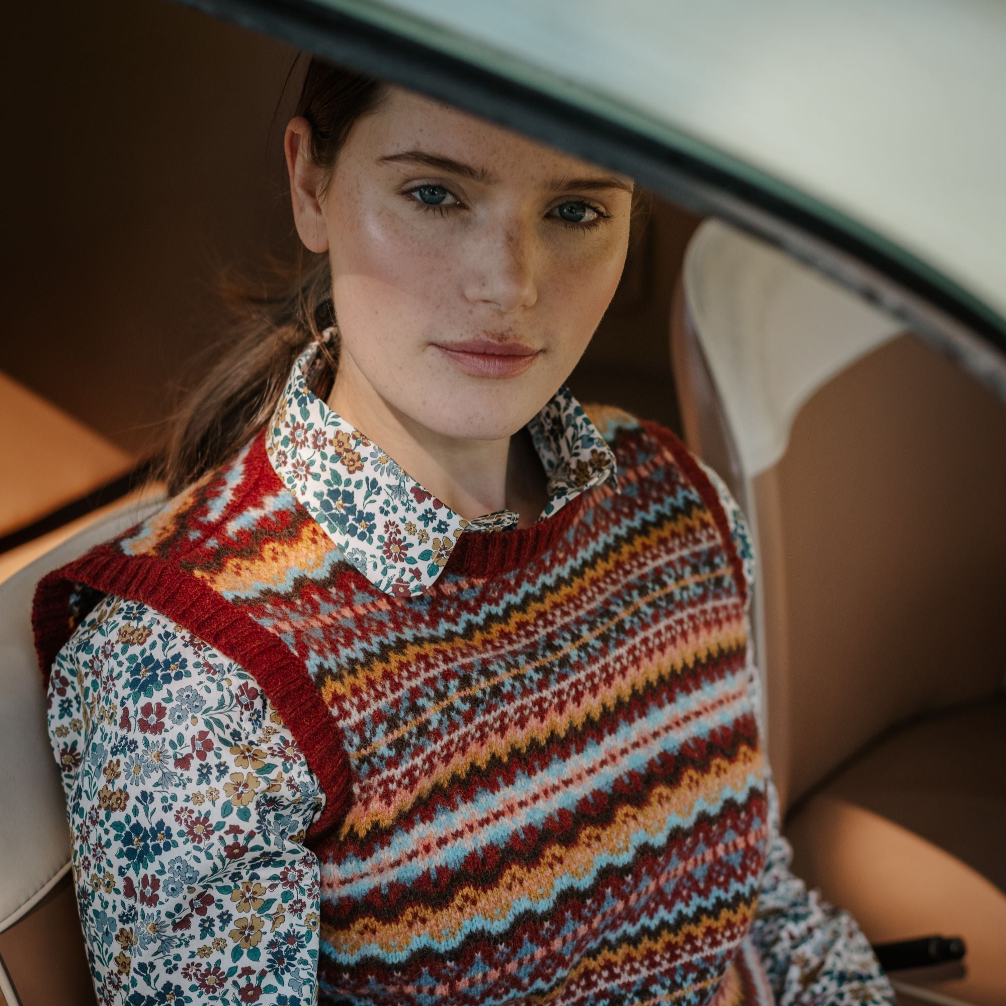 A woman with fair skin and brown hair sits in a car, wearing a colorful patterned knit vest over a floral button-down shirt. She looks calmly at the camera, surrounded by beige car interior.
