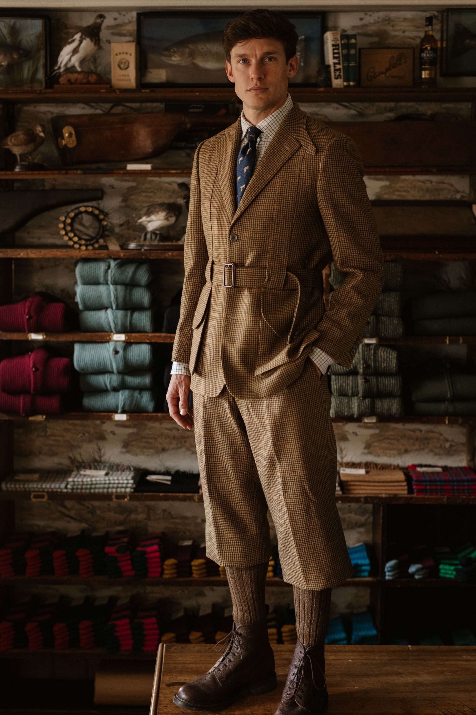 A man stands indoors wearing a vintage-style brown tweed suit with knee-length trousers, long socks, boots, and a tie. Behind him are shelves neatly stacked with folded clothes and accessories in a warmly lit room.
