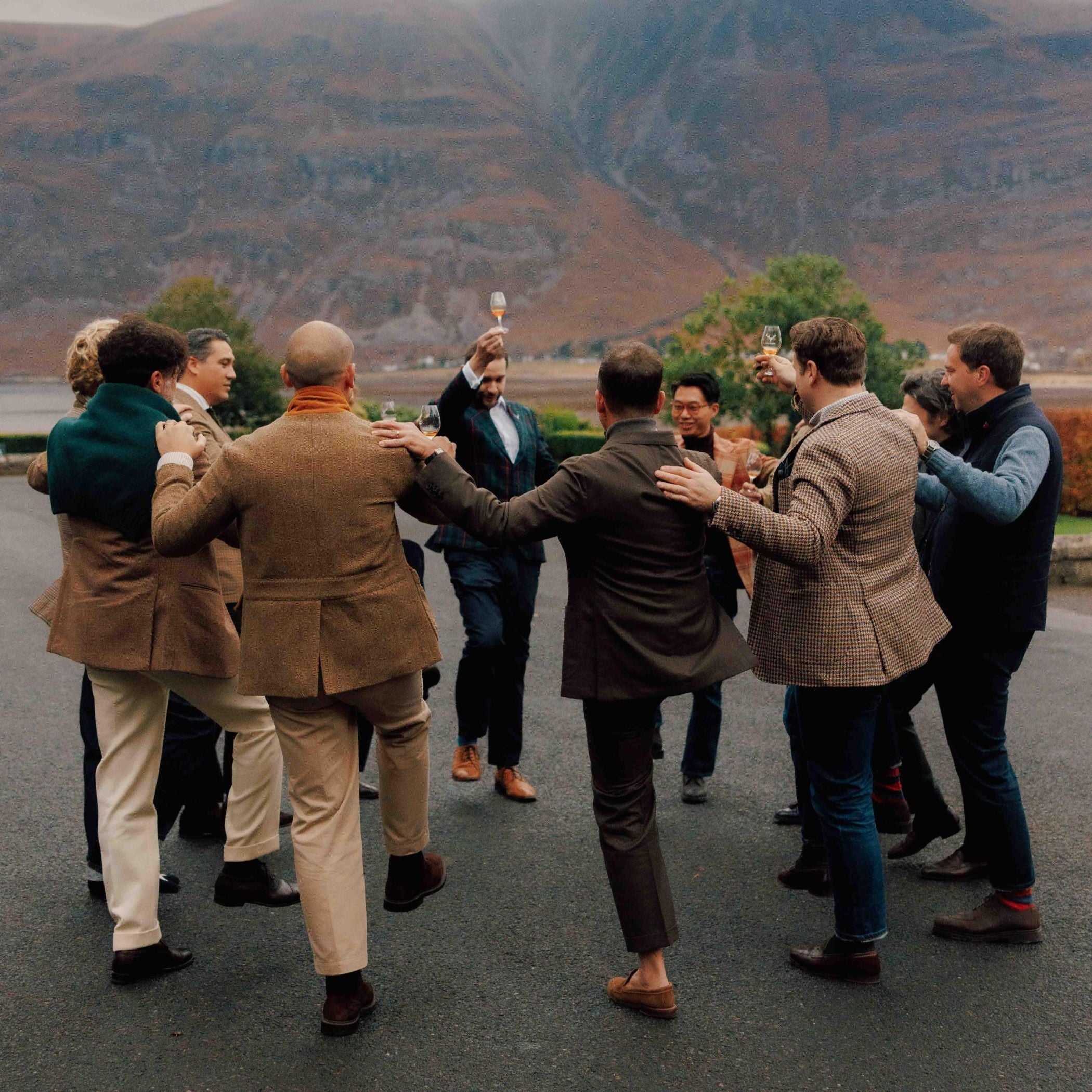 A group of people stand outdoors in a circle, arms linked and legs lifted as they dance together. Mountains and water are visible in the background under an overcast sky.