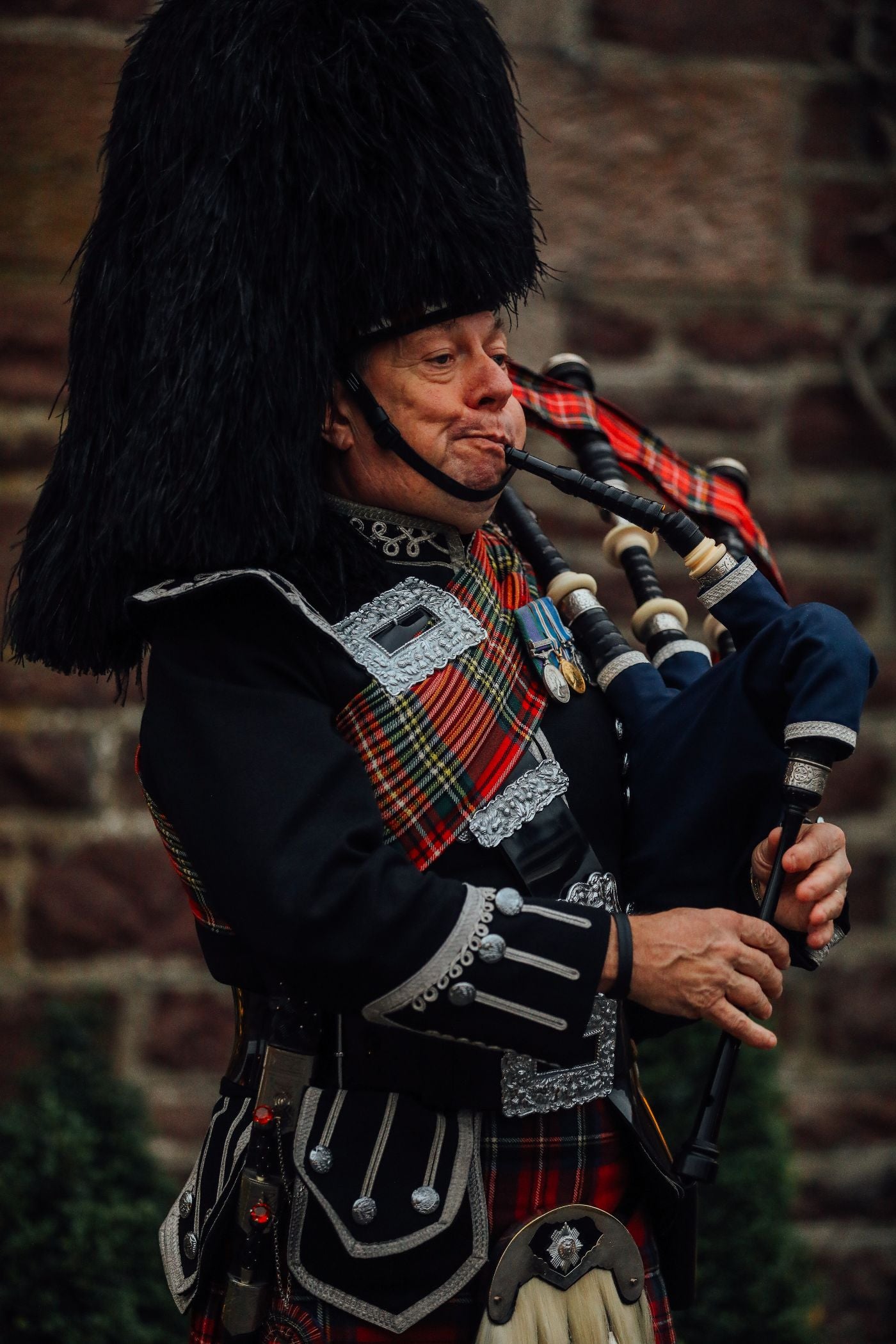 A man wearing traditional Scottish Highland attire, including a feathered hat and tartan sash, plays the bagpipes in front of a stone wall.