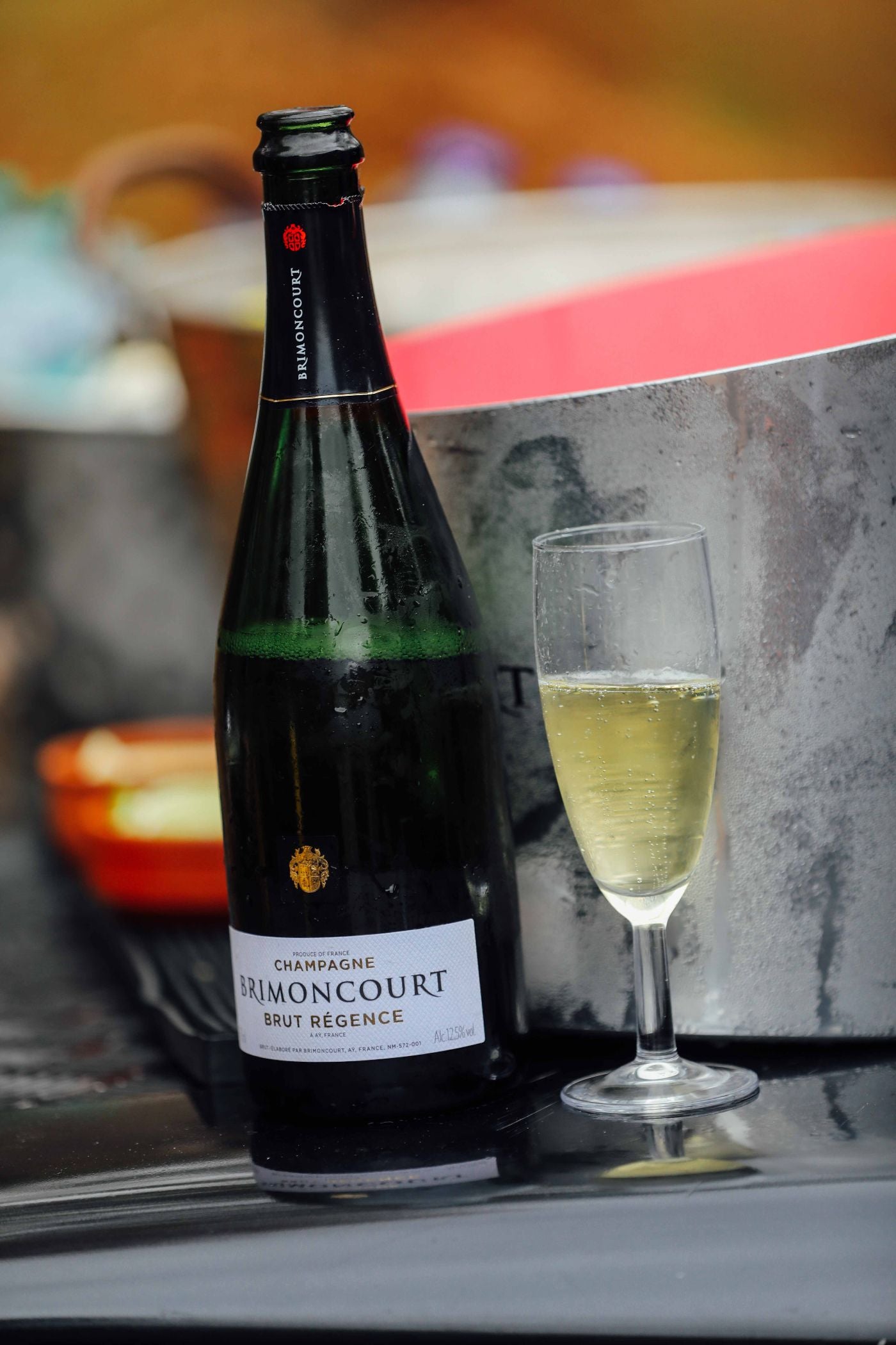 A bottle of Brimoncourt Brut Régence champagne stands next to a filled champagne flute on a reflective surface, with an ice bucket and blurred background visible behind them.