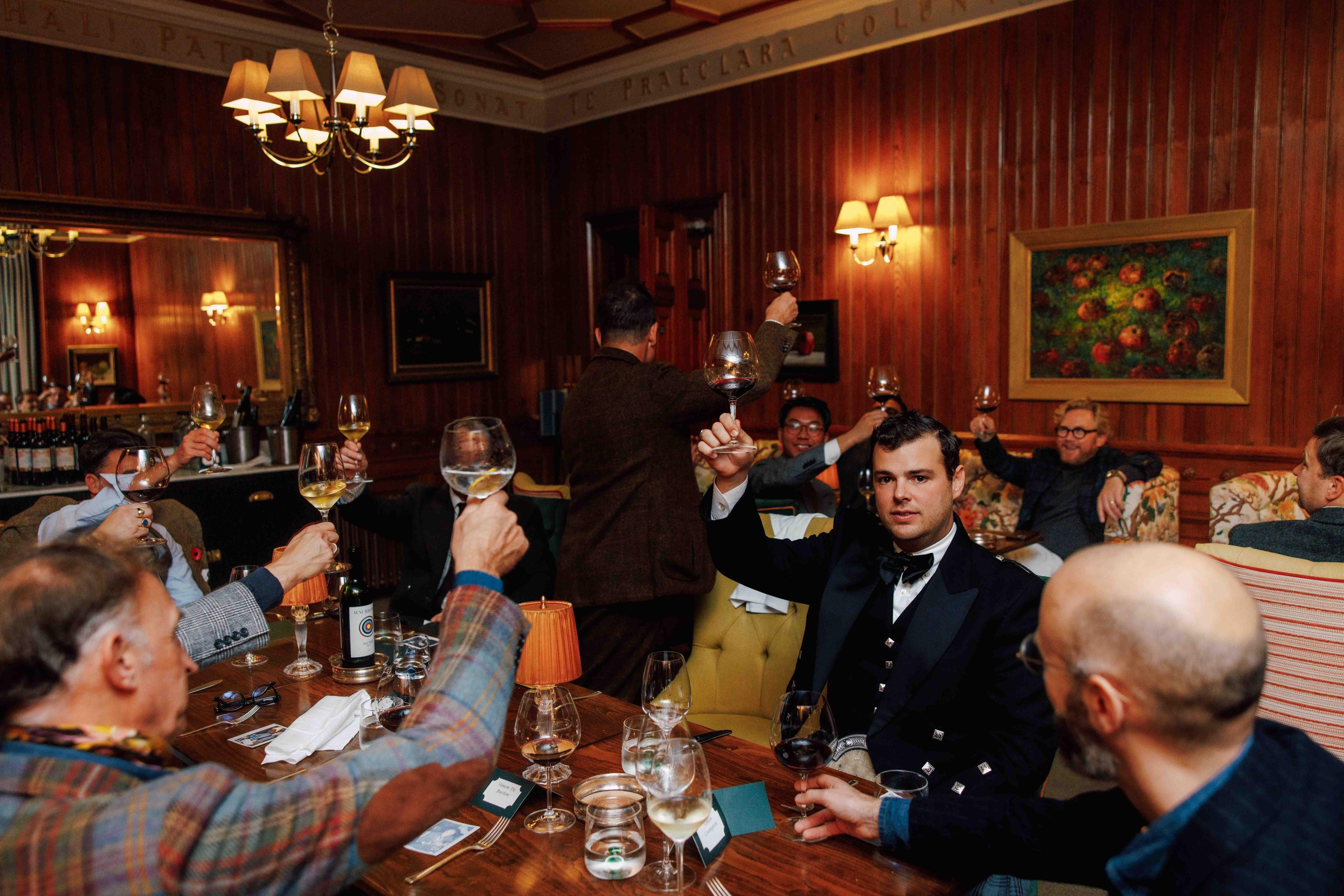 A group of well-dressed people sit around a wooden table in a cozy, wood-paneled room, raising their glasses in a toast. The atmosphere is warm and festive, with wine glasses, bottles, and soft lighting.