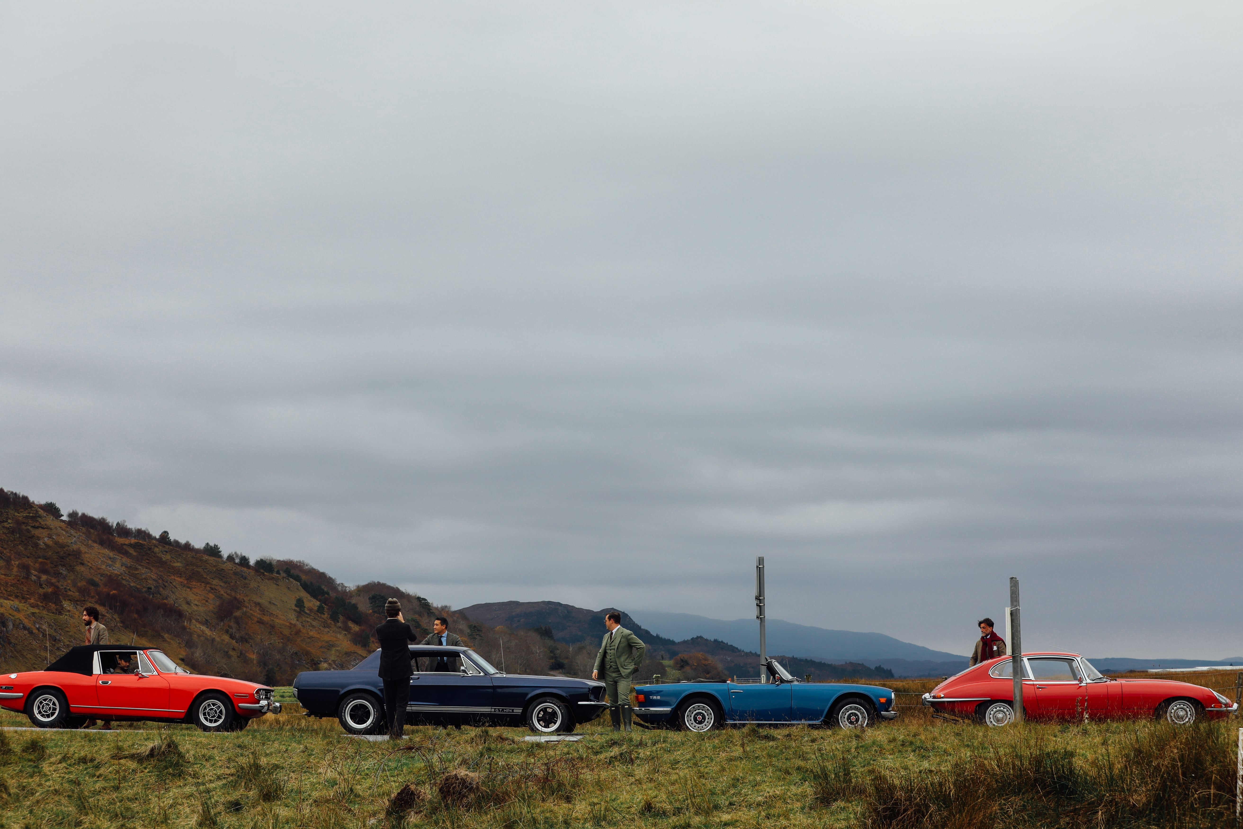 Four people stand beside four classic cars—red, black, blue, and red—parked on grass by a rural road, with hills and a cloudy sky in the background.