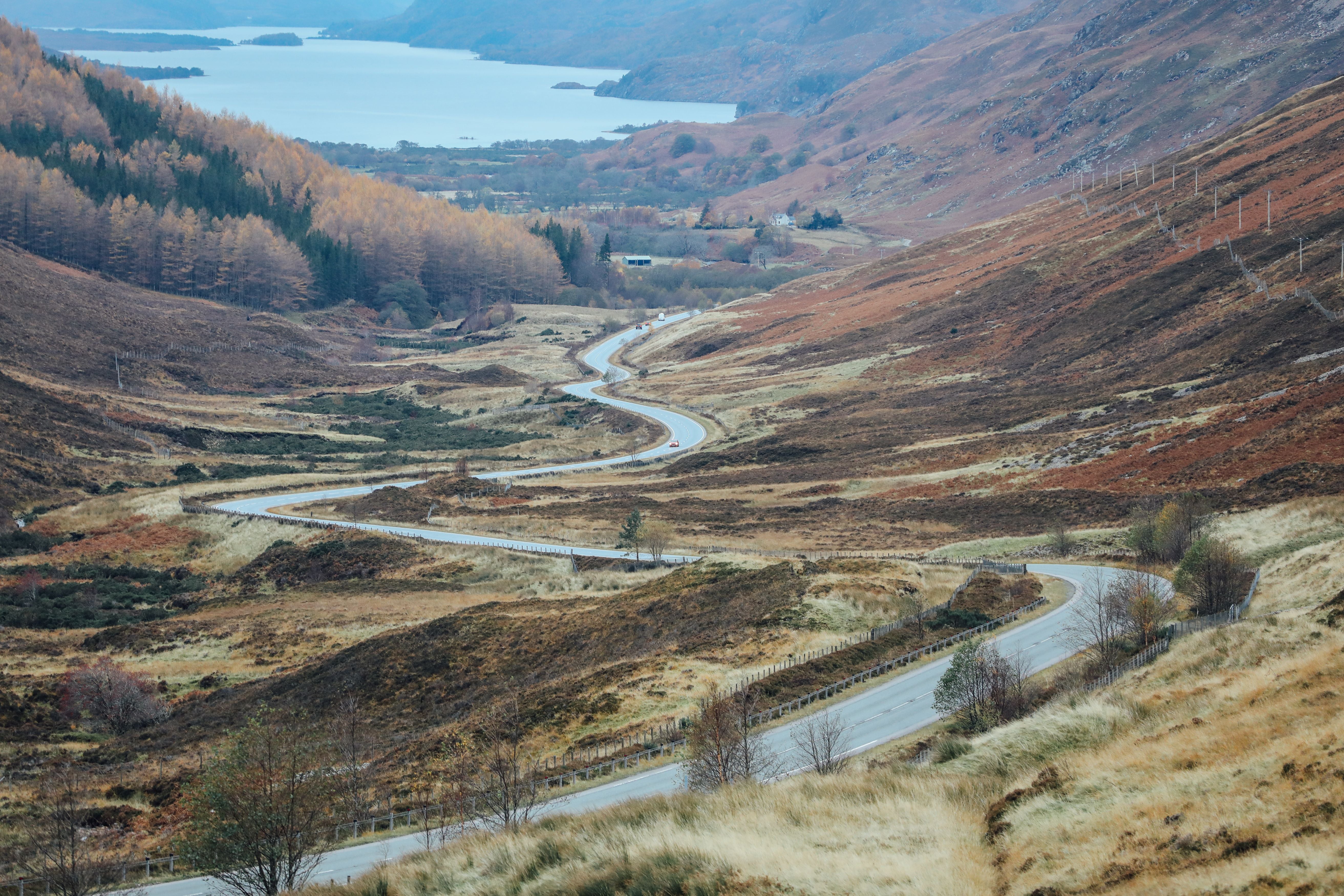 A winding road curves through a hilly, autumn landscape with grass and sparse trees, leading towards a distant lake surrounded by mountains under a cloudy sky.