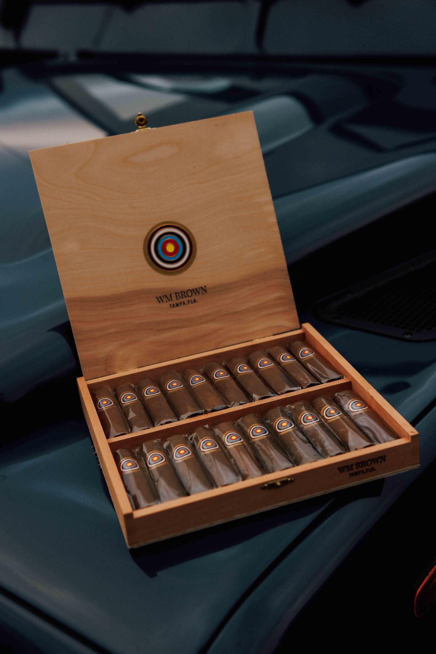 A wooden box with an open lid displays two rows of cigars, each labeled with a round logo. The box rests on the hood of a dark blue car. The inside of the lid reads WM BROWN and ZAMPA 7.14.