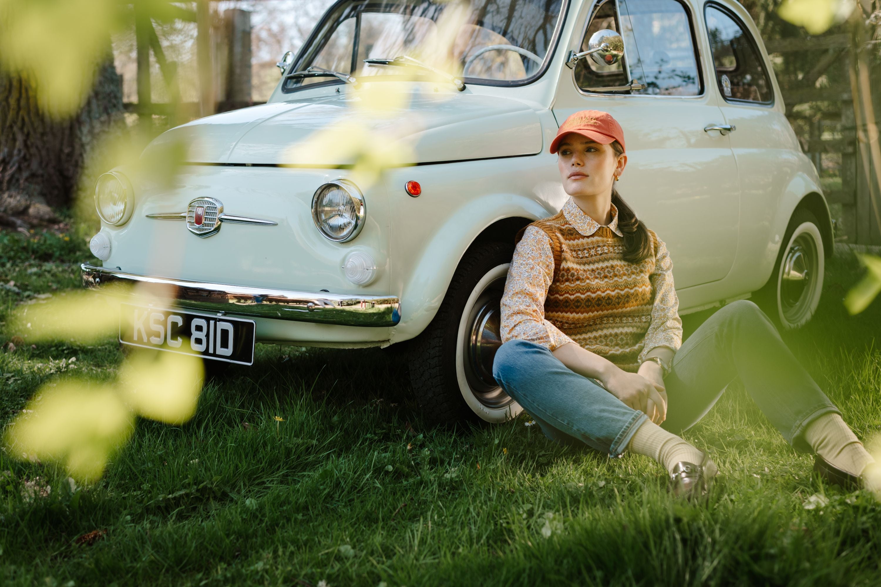 A woman in casual vintage-style clothing and a cap sits on grass, leaning against a classic white Fiat car with UK plates, surrounded by green foliage on a sunny day.