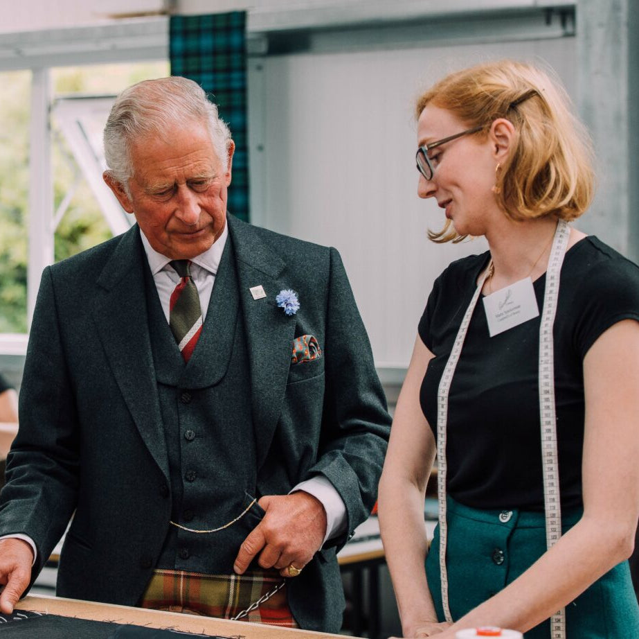An older man in a suit with a flower on his lapel stands beside a woman wearing glasses and a measuring tape around her neck. They are indoors, talking and looking at fabric on a table.