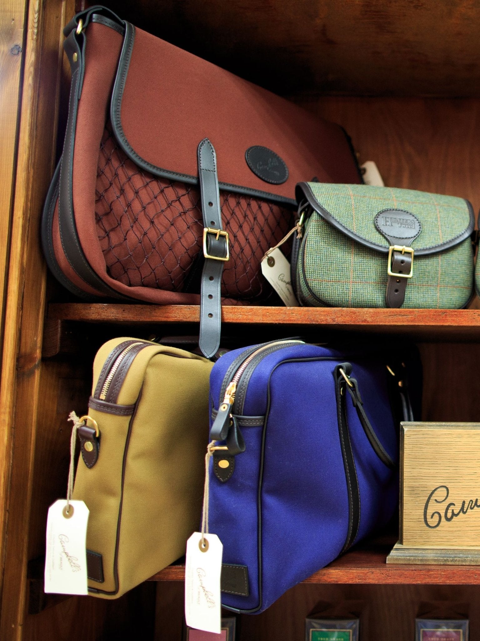 Two wooden shelves showcase Campbell's of Beauly bags: a large brown padded Laptop Bag and a green crossbody on the top shelf, with tan and blue duffel bags below. All have leather details and tags, ideal for work or travel.