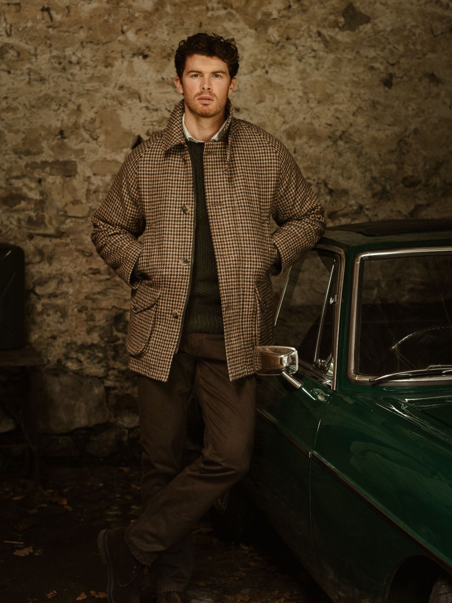 A man in Campbell's of Beauly Cannich Coat—a brown plaid country coat with storm collar—stands, hands in pockets, by a vintage green car against a stone wall in a rustic garage, wearing dark pants.