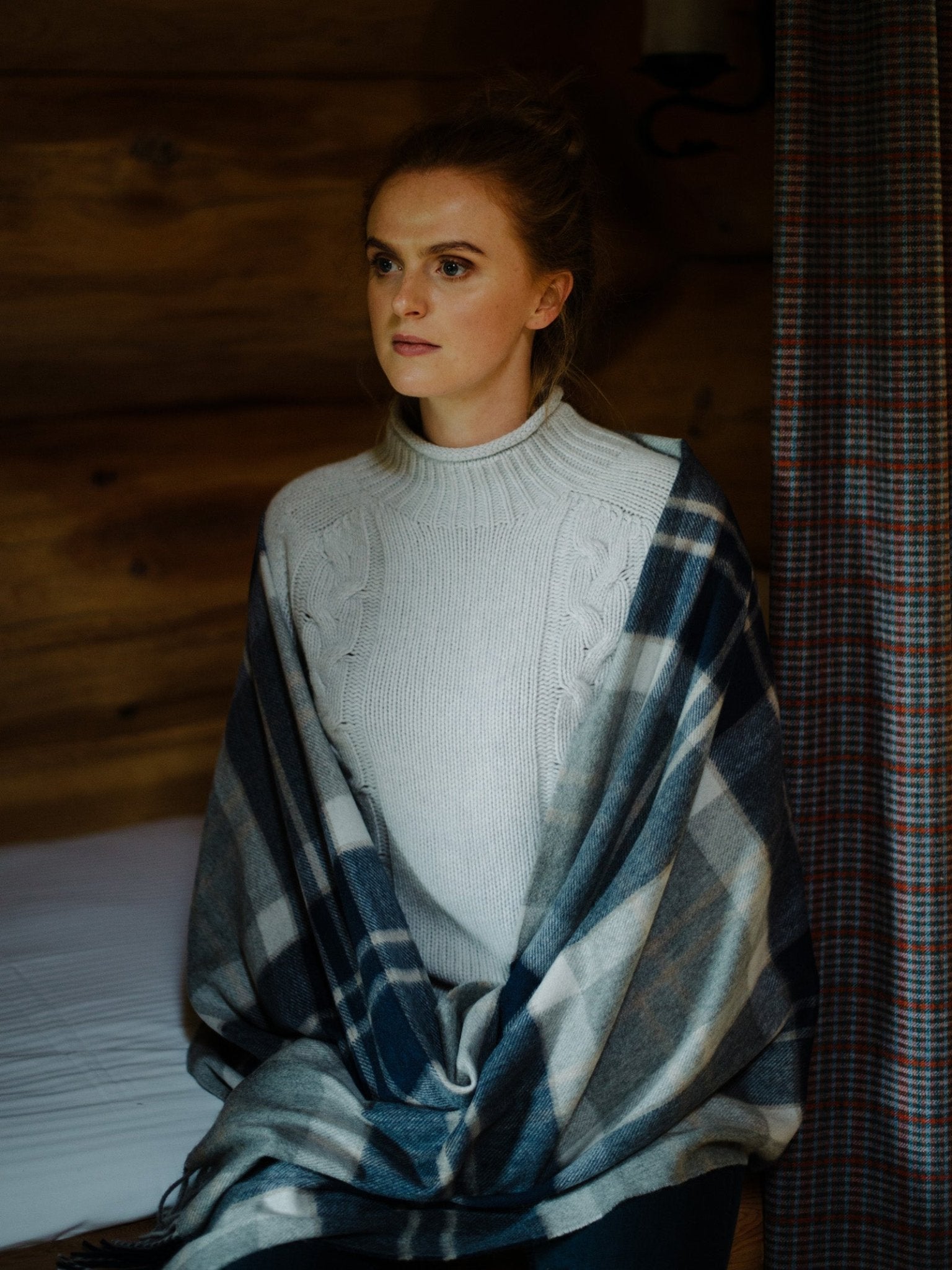 A woman with light brown hair, wearing a white knit sweater and a blue plaid Campbell's of Beauly Cashmere Stole, sits on a bed indoors by a wooden wall and plaid curtain, gazing calmly to the side.