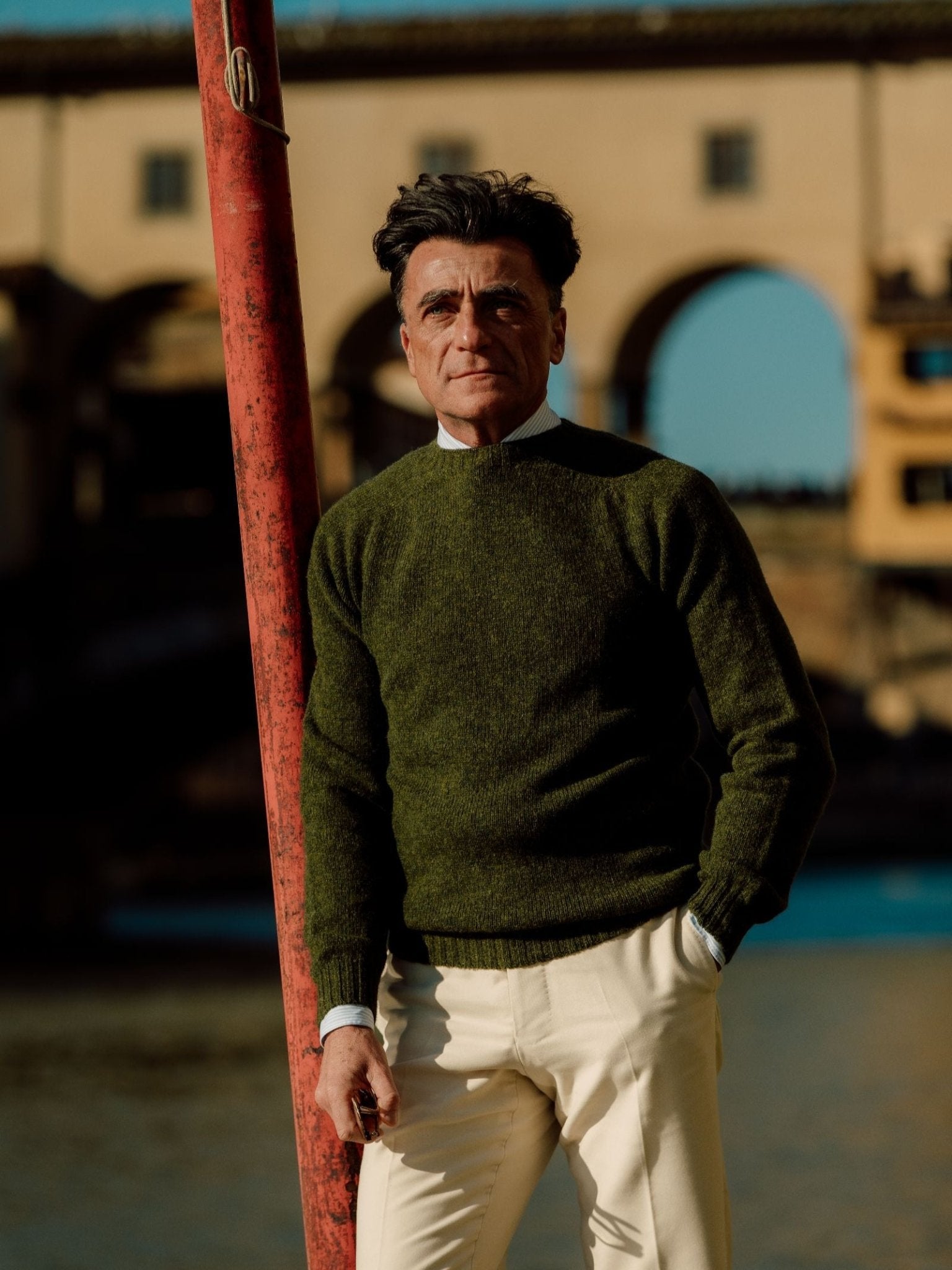A man wearing the Campbell's of Beauly Shetland Jumper stands outdoors in beige pants, leaning against a red pole, with a historic stone bridge and blue sky in the background.