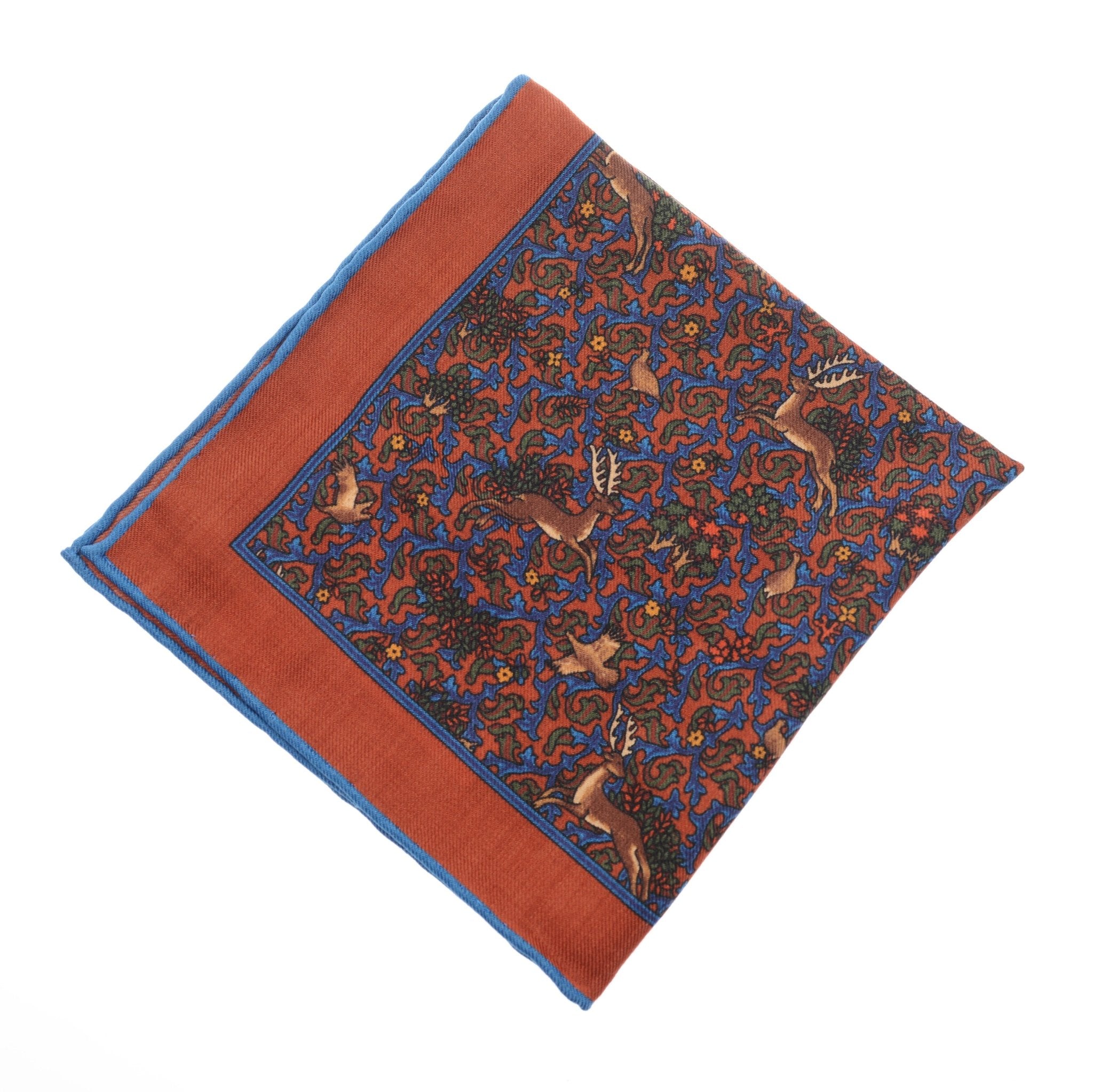 The Wool Hank by Campbells of Beauly is a red pocket square with hand-rolled edges, featuring a central deer motif and elegant blue and red scrolling, making it a timeless accessory.