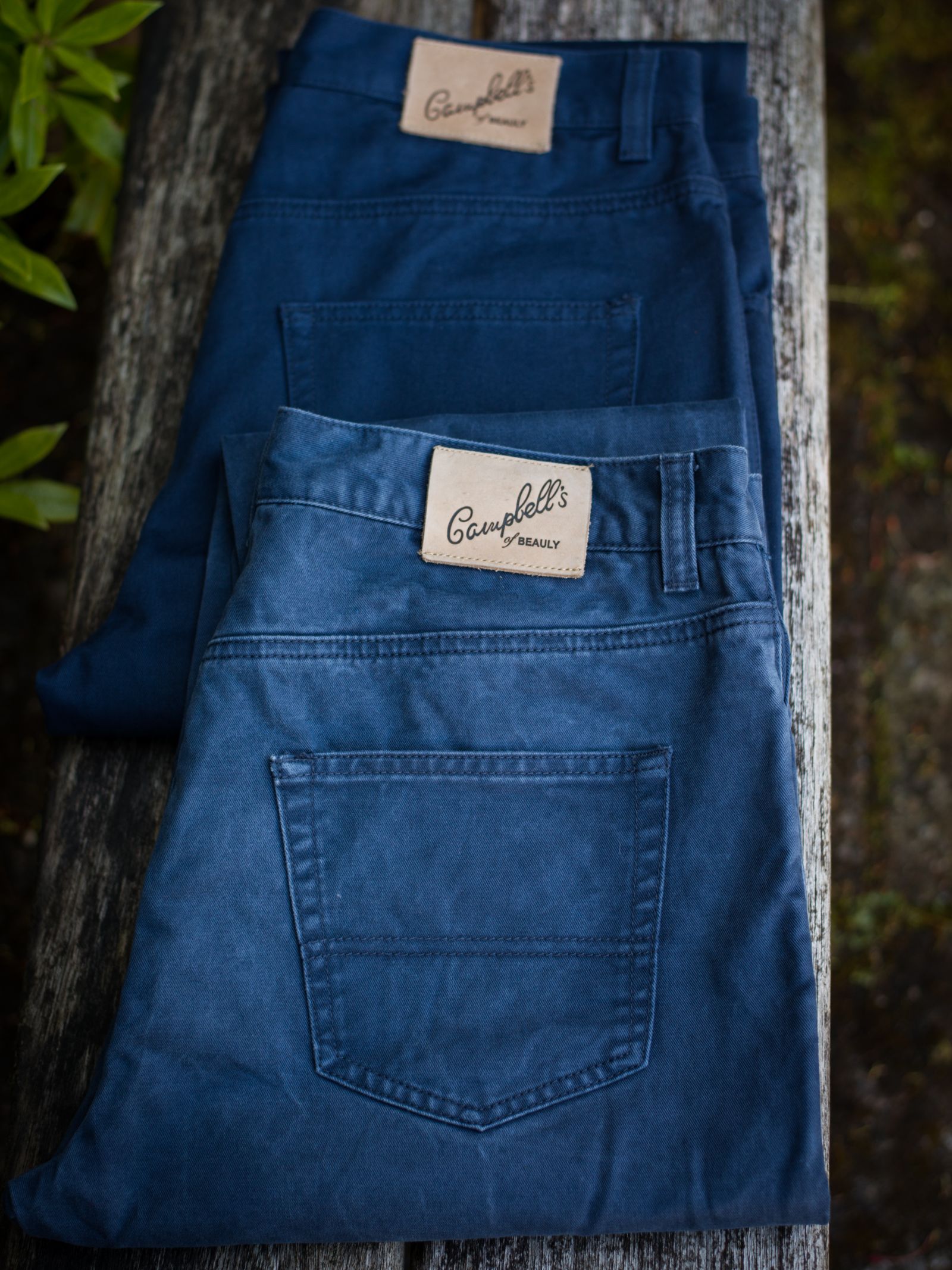 Two pairs of Campbell's of Beauly Colour Cotton Jeans, straight leg in blue, classic five-pocket style, folded on a weathered wooden surface—one pair slightly darker than the other.