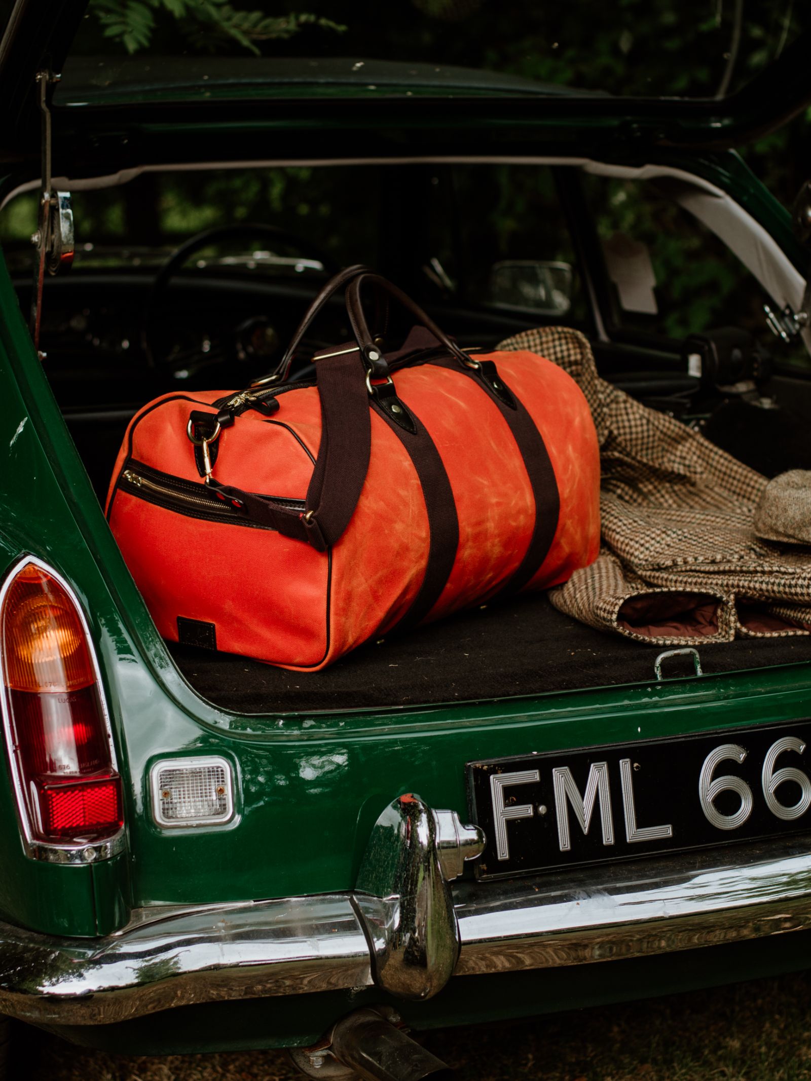 The Campbell's of Beauly Wax Holdall, featuring bright orange waxed canvas with dark straps and bridle leather handles, rests in the open trunk of a vintage green car beside a checkered jacket. The license plate reads “FML 66.”.