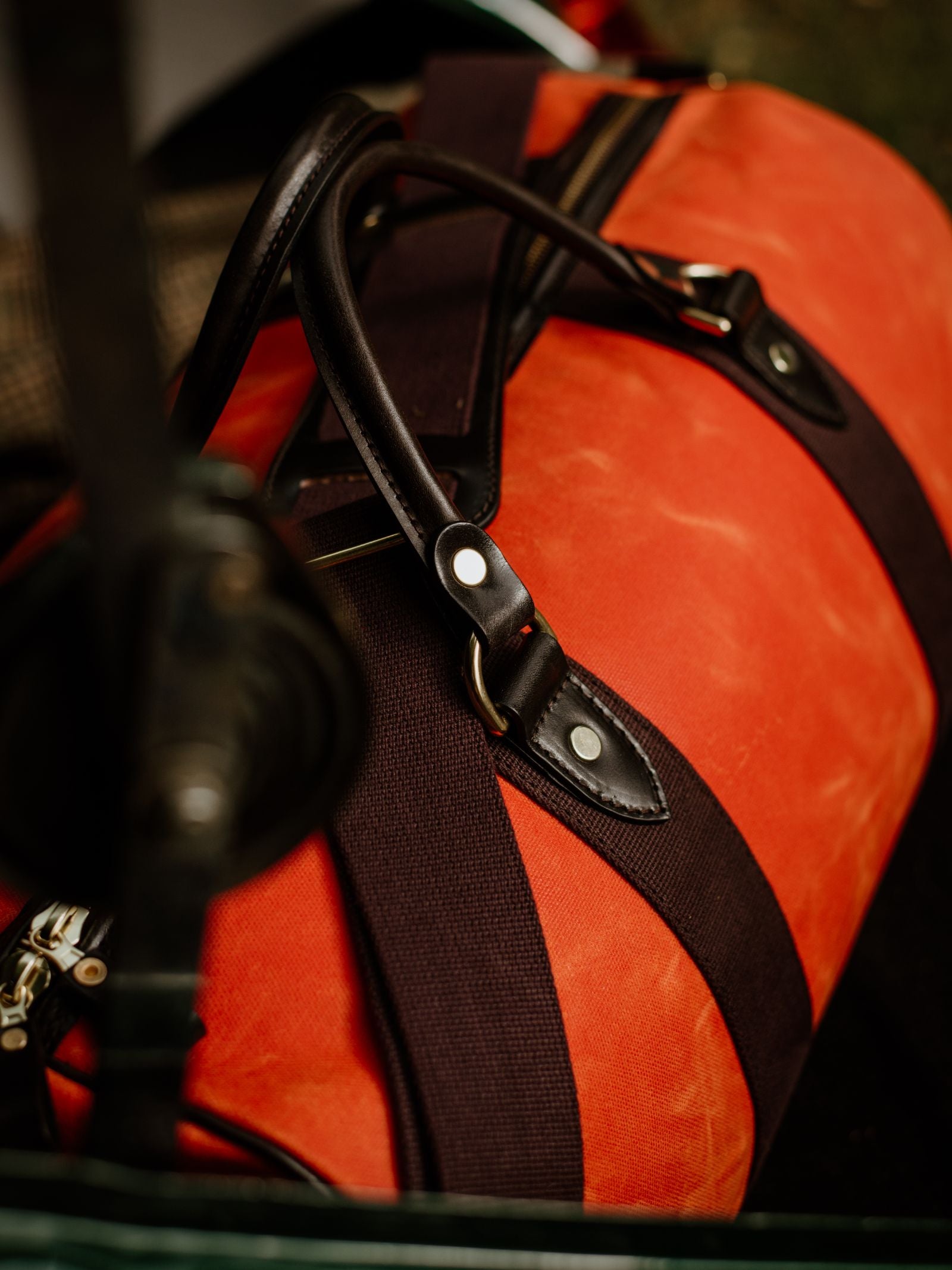 A close-up of the Campbell's of Beauly Wax Holdall, featuring orange waxed canvas, black straps, and bridle leather handles, placed on a surface with blurred background objects.