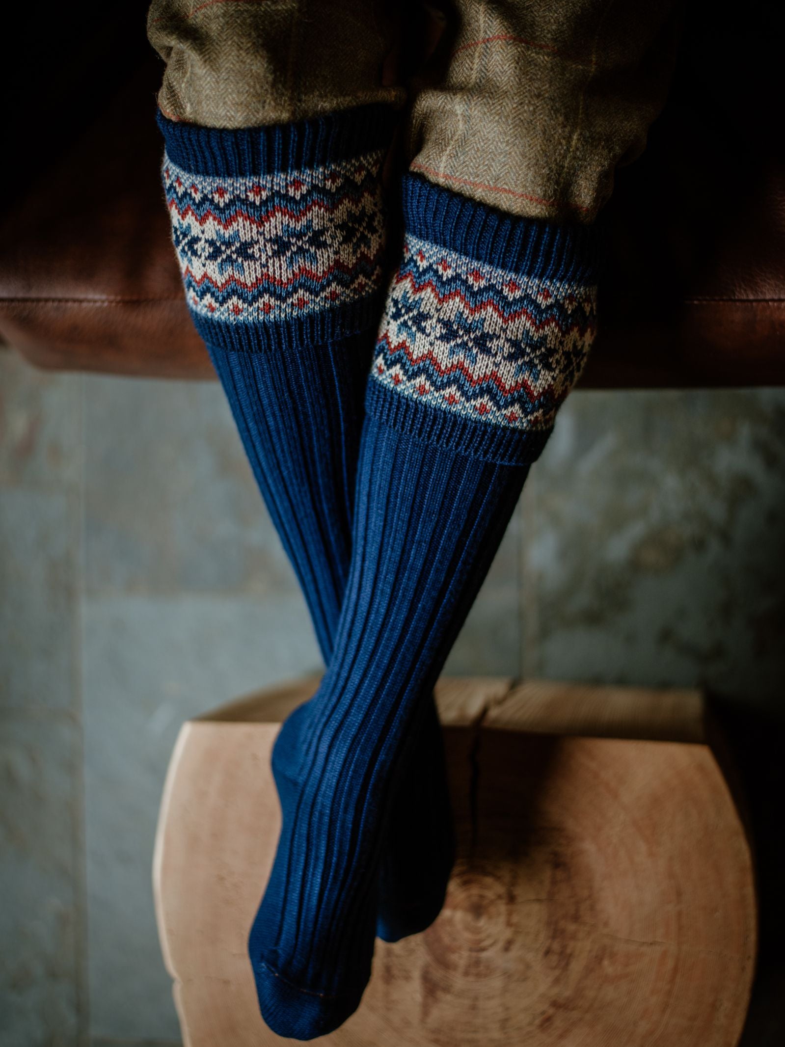 Close-up of legs wearing Campbell's of Beauly Merino Fairisle Shooting Socks—dark blue with a patterned red, cream, and blue cuff. Paired with brown tweed trousers, the look highlights traditional Scottish knitwear.