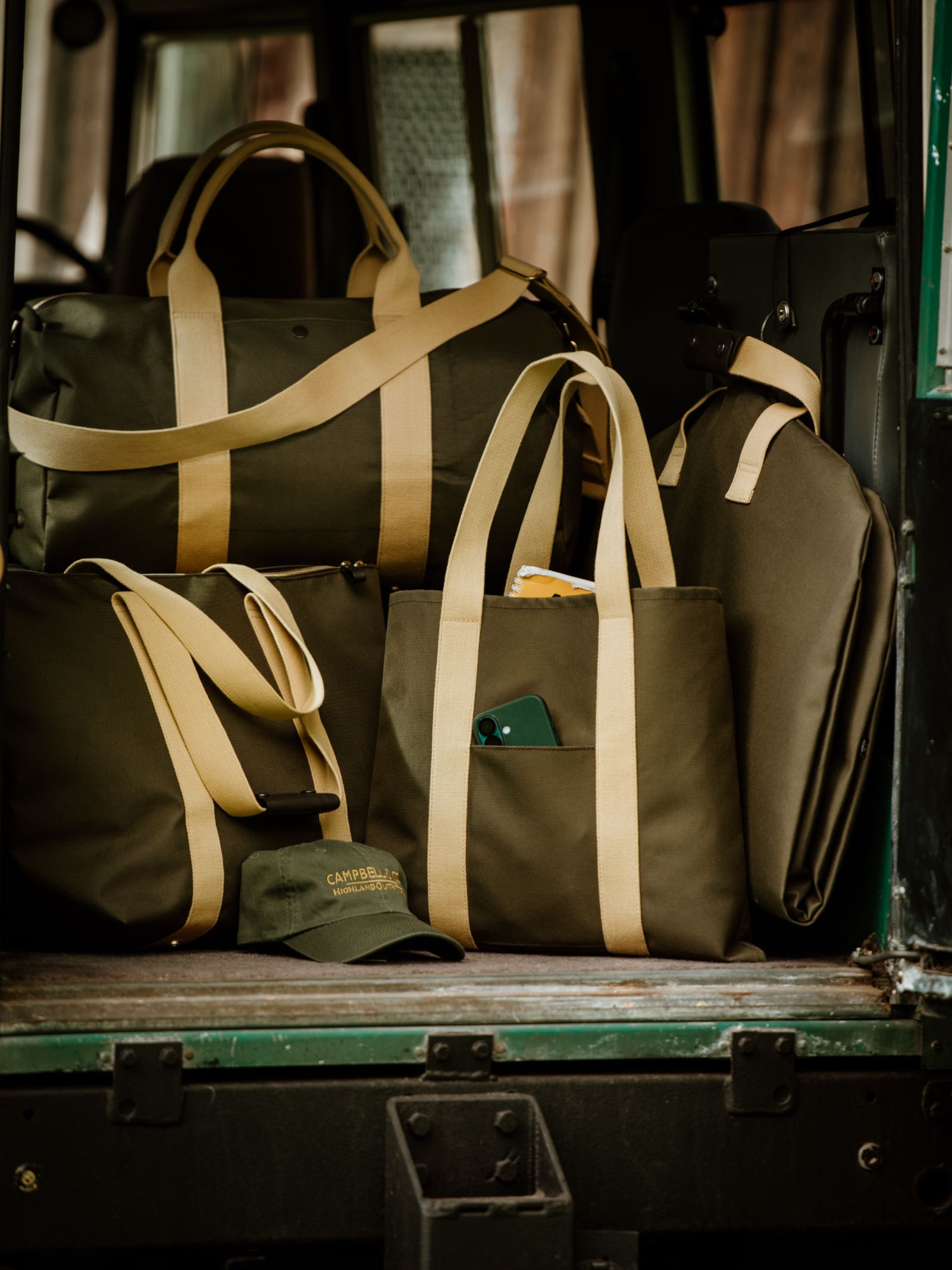 Several olive-green Utility Medium Totes by Campbell's of Beauly with tan straps are arranged in a vehicle, alongside a matching canvas tote. A green CAPRIVI cap is placed in front, making the setup ideal for everyday carry.