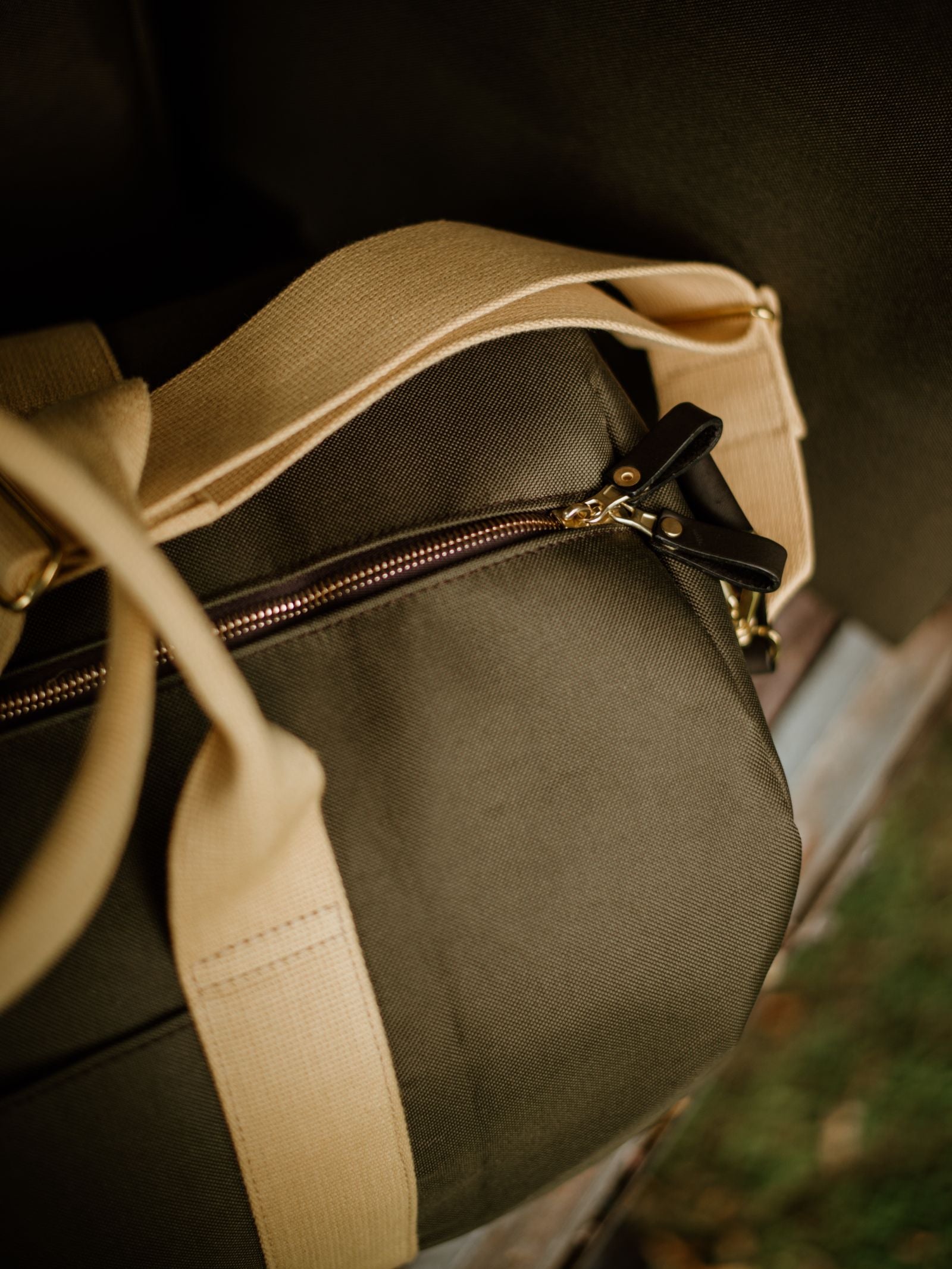 A close-up of the Utility Medium Duffle by Campbell's of Beauly, a durable olive green travel bag with beige straps and a gold zipper, photographed outdoors with a blurred green background.