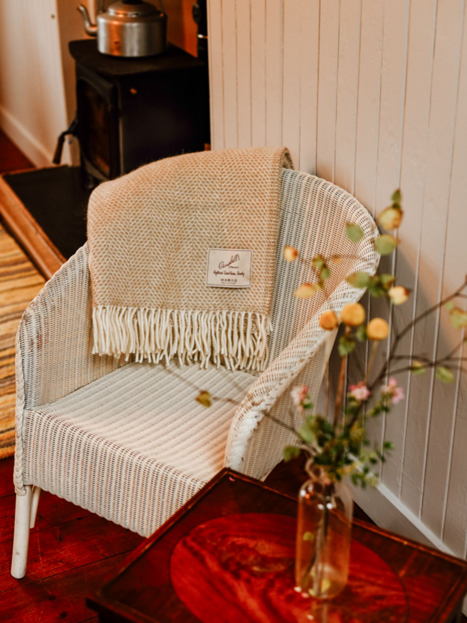 A wicker chair with a breathable weave features a Campbell's of Beauly Stipled Twill Blanket draped over it, beside a wooden side table with flowers, near a wood stove and a white paneled wall.
