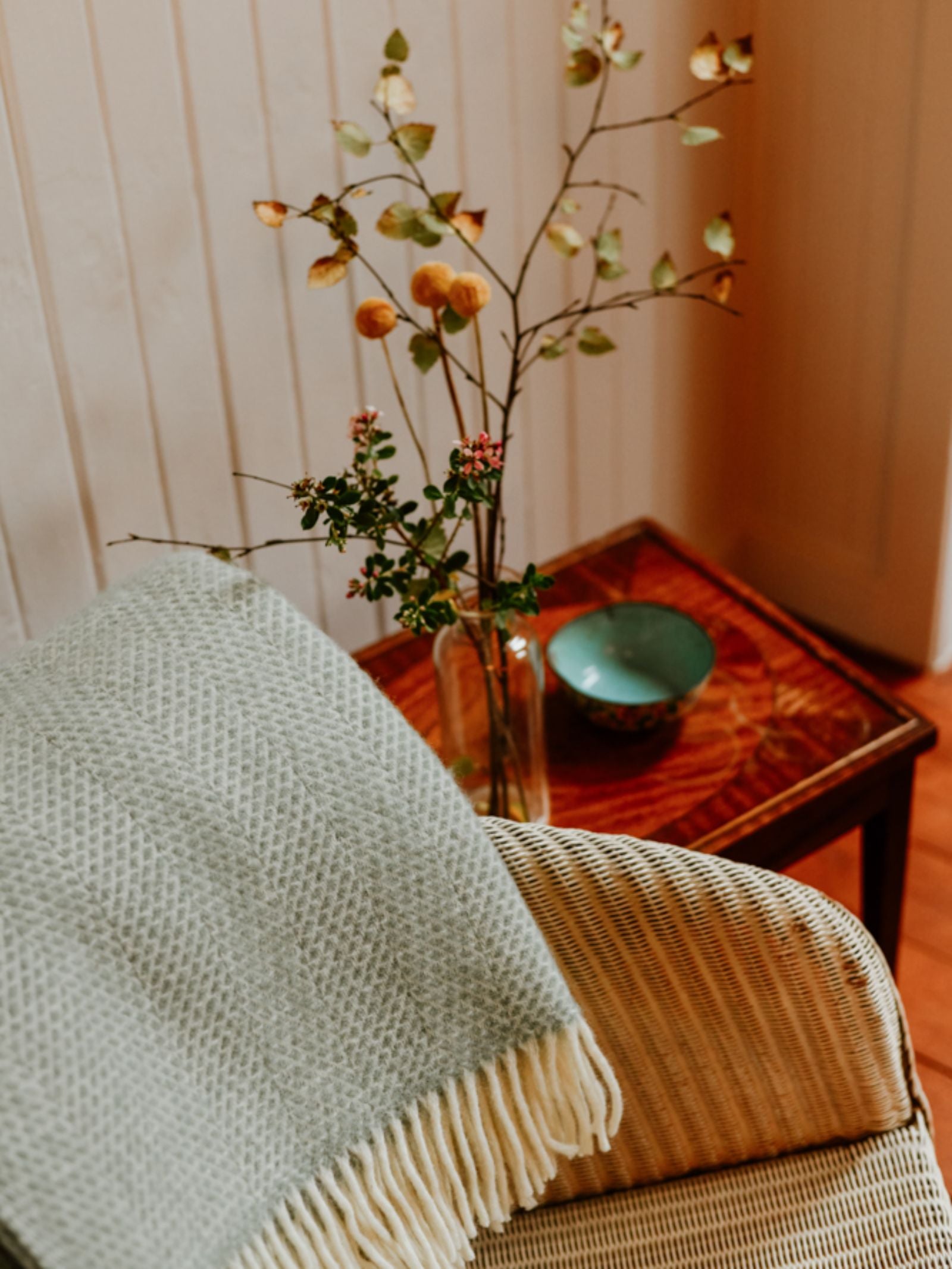 A cozy wicker chair with a Campbell's of Beauly Stipled Twill Blanket in pale green draped over it sits beside a wooden side table topped with a vase of autumn branches and a decorative bowl.