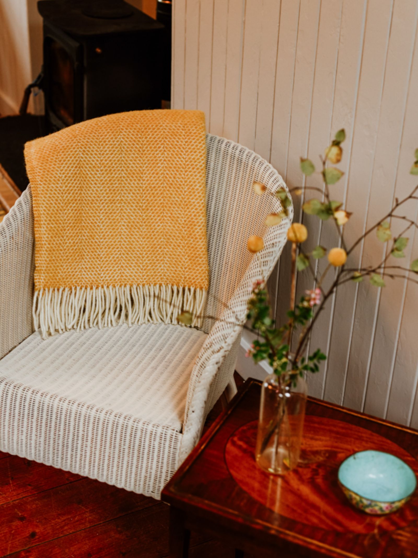 A white wicker chair with a Campbell's of Beauly Stipled Twill Blanket draped over the back sits next to a wooden side table holding a glass vase with branches and a small turquoise bowl.