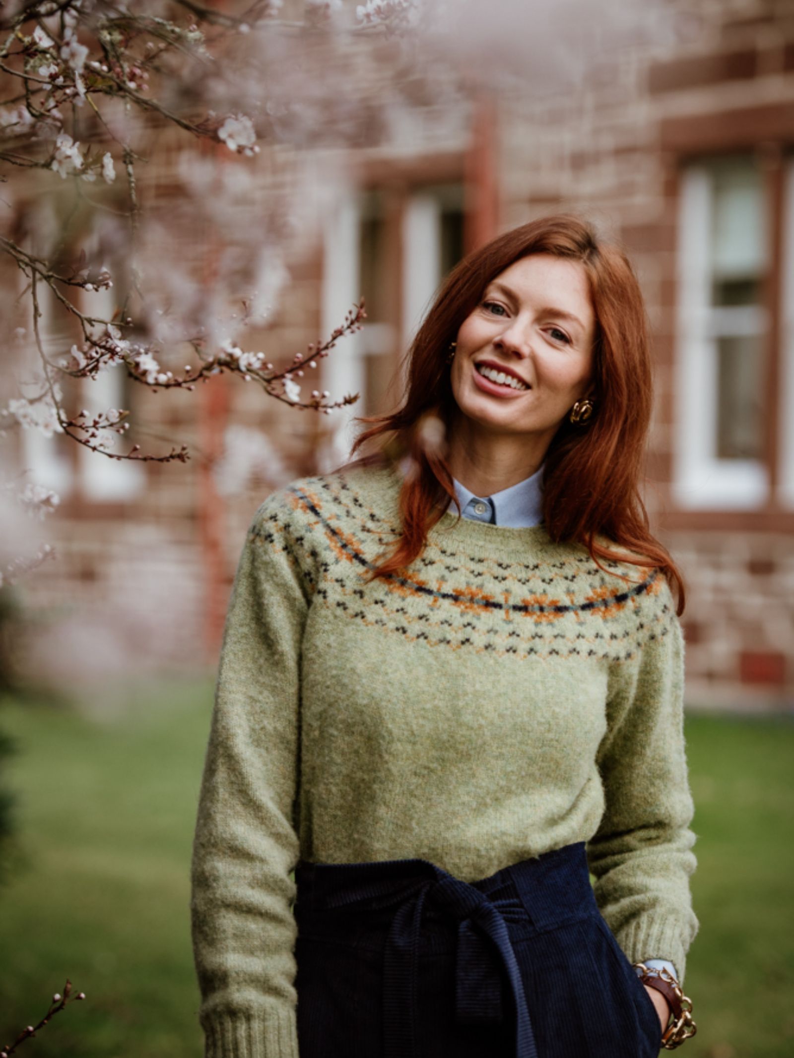 A woman with long red hair smiles outdoors near blossoming branches, wearing a light green Auld Stock Cropped Fairisle Crew Neck Jumper by Campbell's of Beauly over a collared shirt and dark skirt. A stone building with windows is behind her.