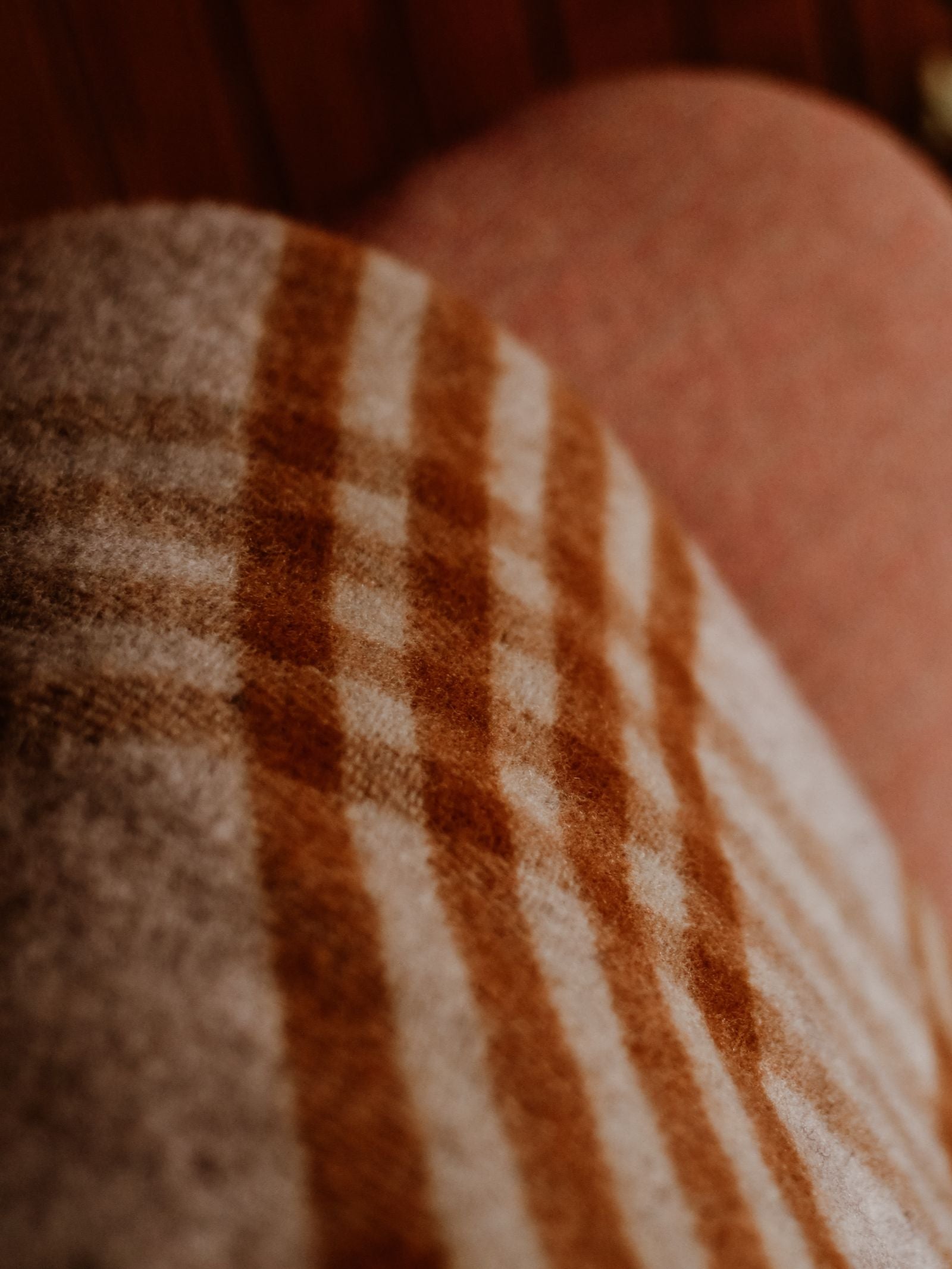 The Tarradale Lambswool Throw by Campbell's of Beauly showcases a soft brown and beige check pattern, elegantly draped with a warm-toned, blurred background.