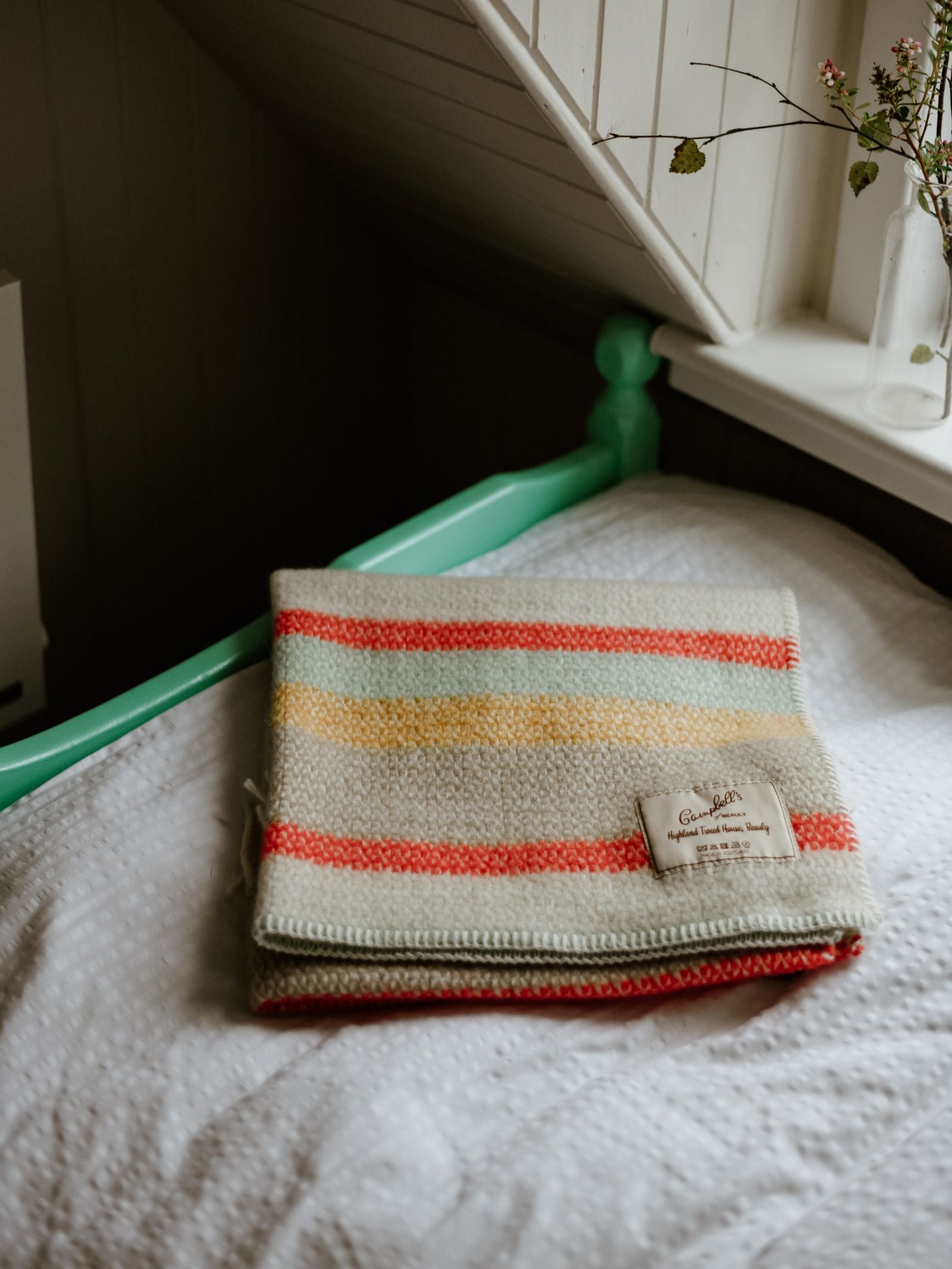 A folded Campbell's of Beauly Baby Blanket in red, yellow, and cream stripes rests on a white textured bedspread beside a mint green bed frame under a slanted ceiling, near a window with a vase holding a twig.