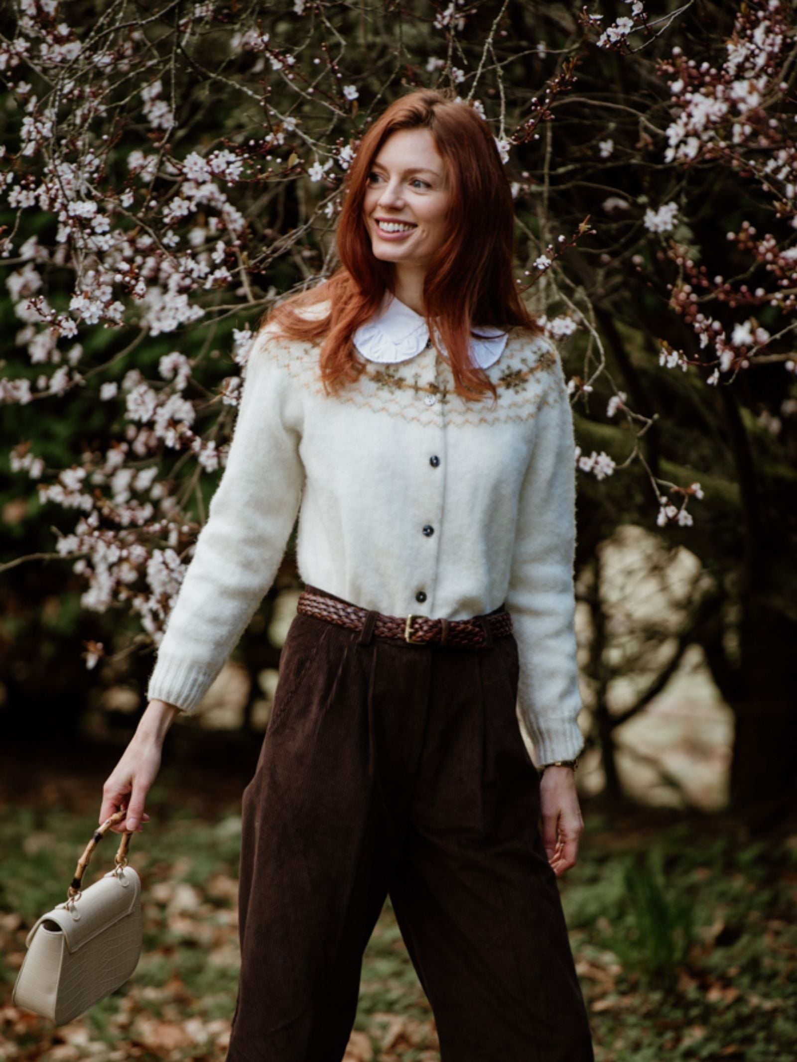 A woman with long red hair smiles outdoors before a blossoming tree. She wears a white sweater with a decorative collar and Campbell's of Beauly Auld Stock Wide Leg Cords in Chocolate, holding a cream-colored handbag.