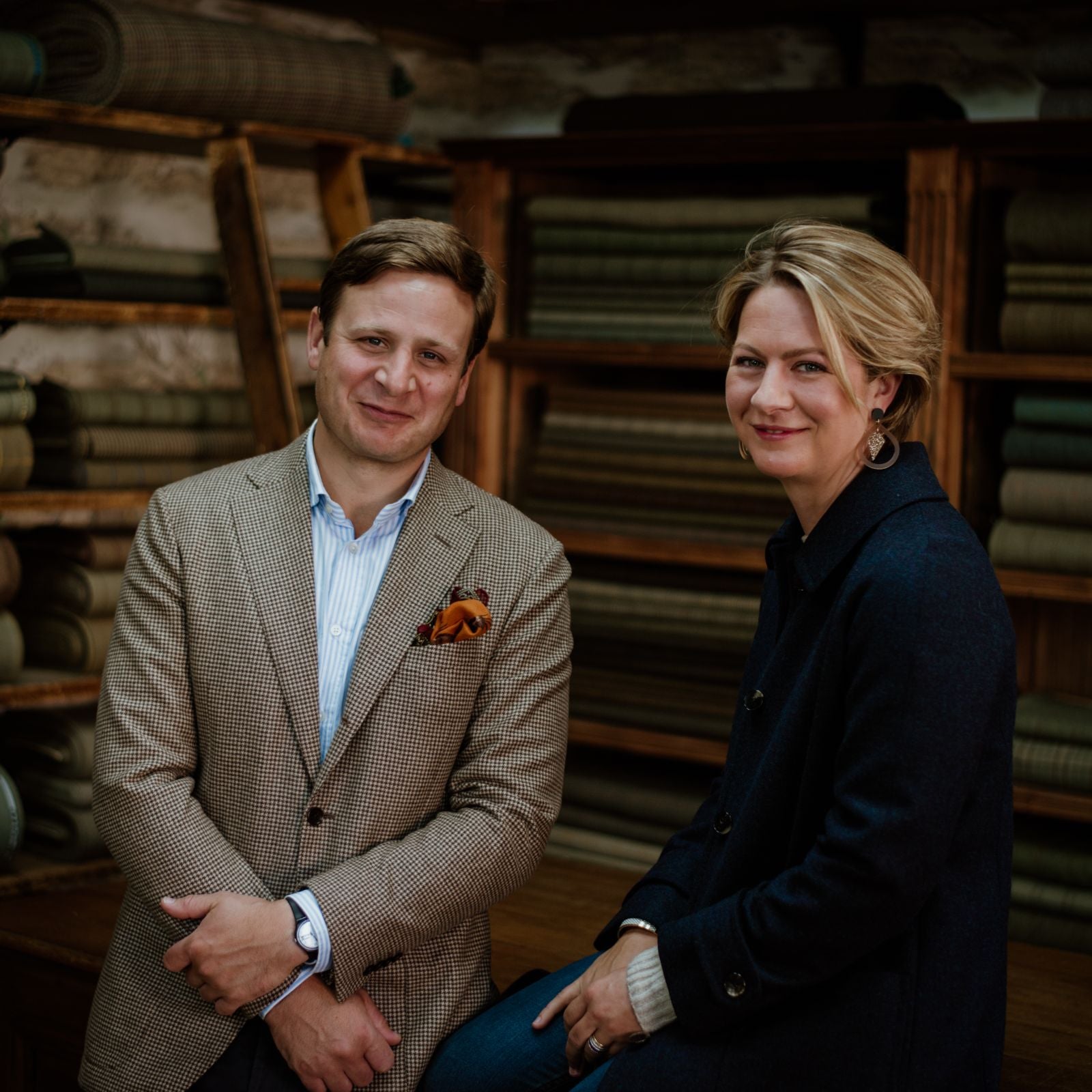 A man in a beige checked blazer and a woman in a dark coat sit and smile in front of shelves stacked with folded fabrics in a warmly lit room.