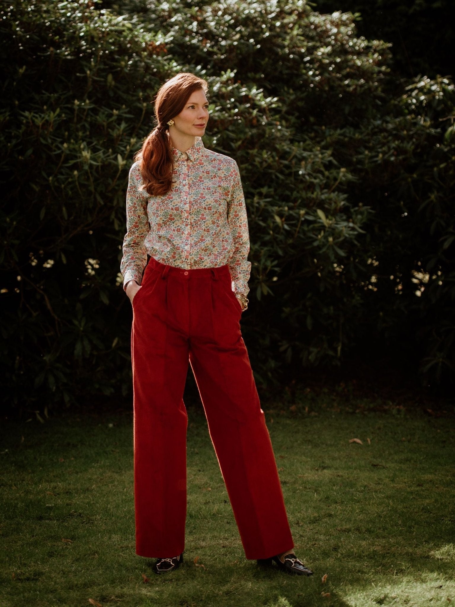 A person with long red hair stands on grass in front of bushes, wearing a floral shirt, dark shoes, and rust Auld Stock Wide Leg Cords by Campbell's of Beauly. They look to the side with hands in pockets.
