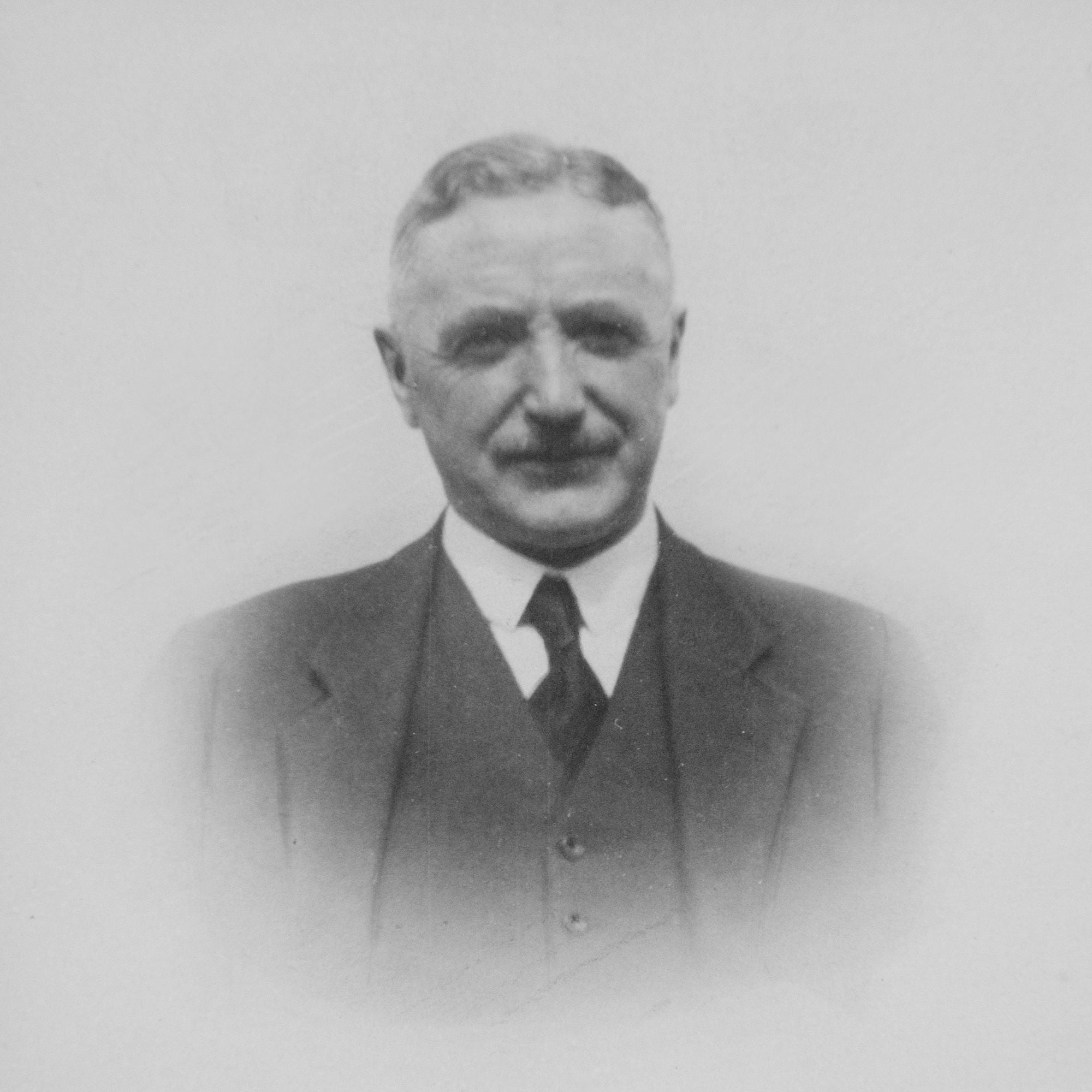 Black and white portrait of an older man with short hair and a mustache, wearing a suit, tie, and vest, facing the camera with a neutral expression against a plain background.