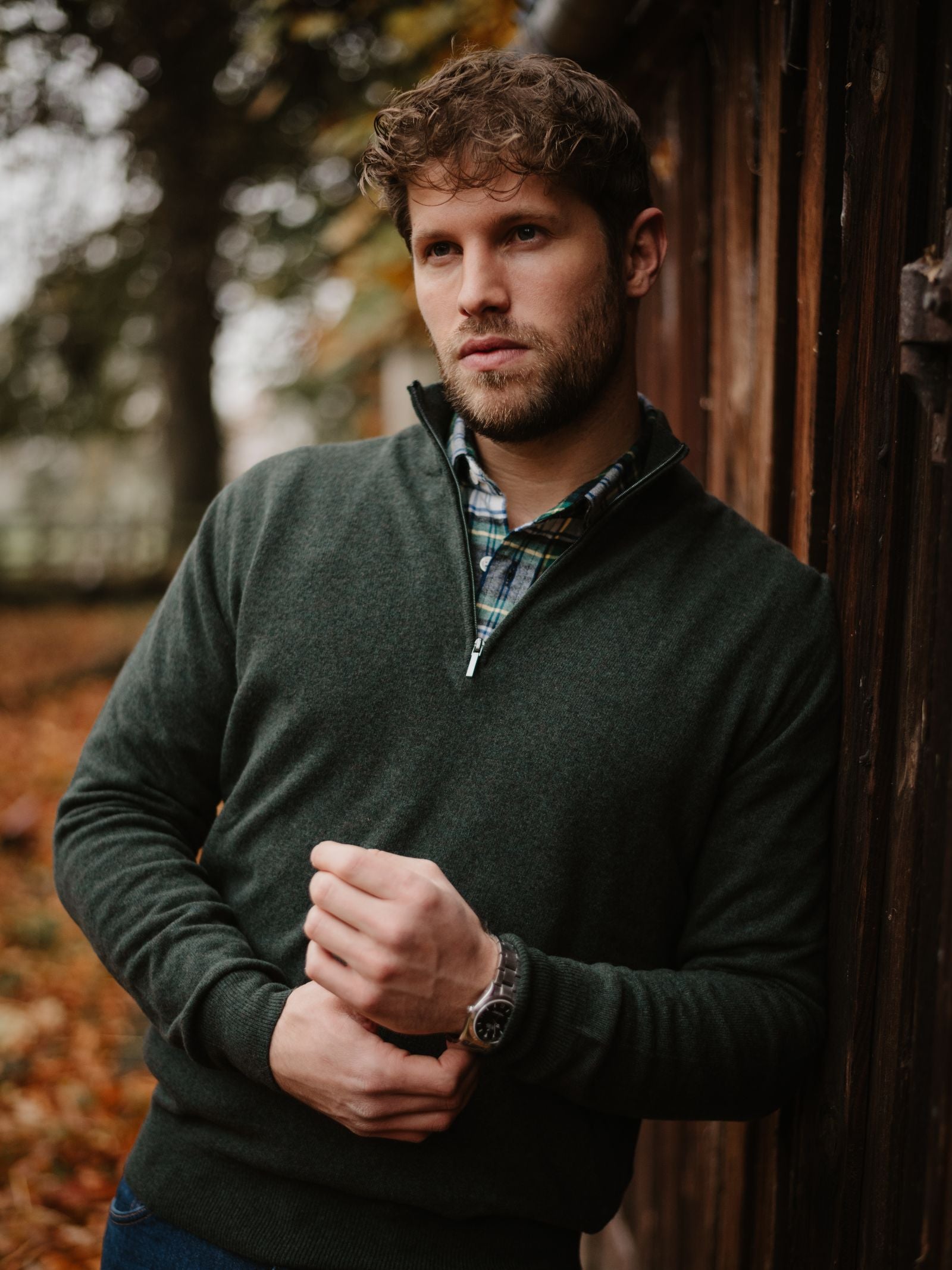 A man with wavy hair and a beard wears the Campbell's of Beauly Merino & Cashmere Quarter Zip Jumper over a plaid shirt, standing outdoors by a wooden structure amid autumn leaves and trees.