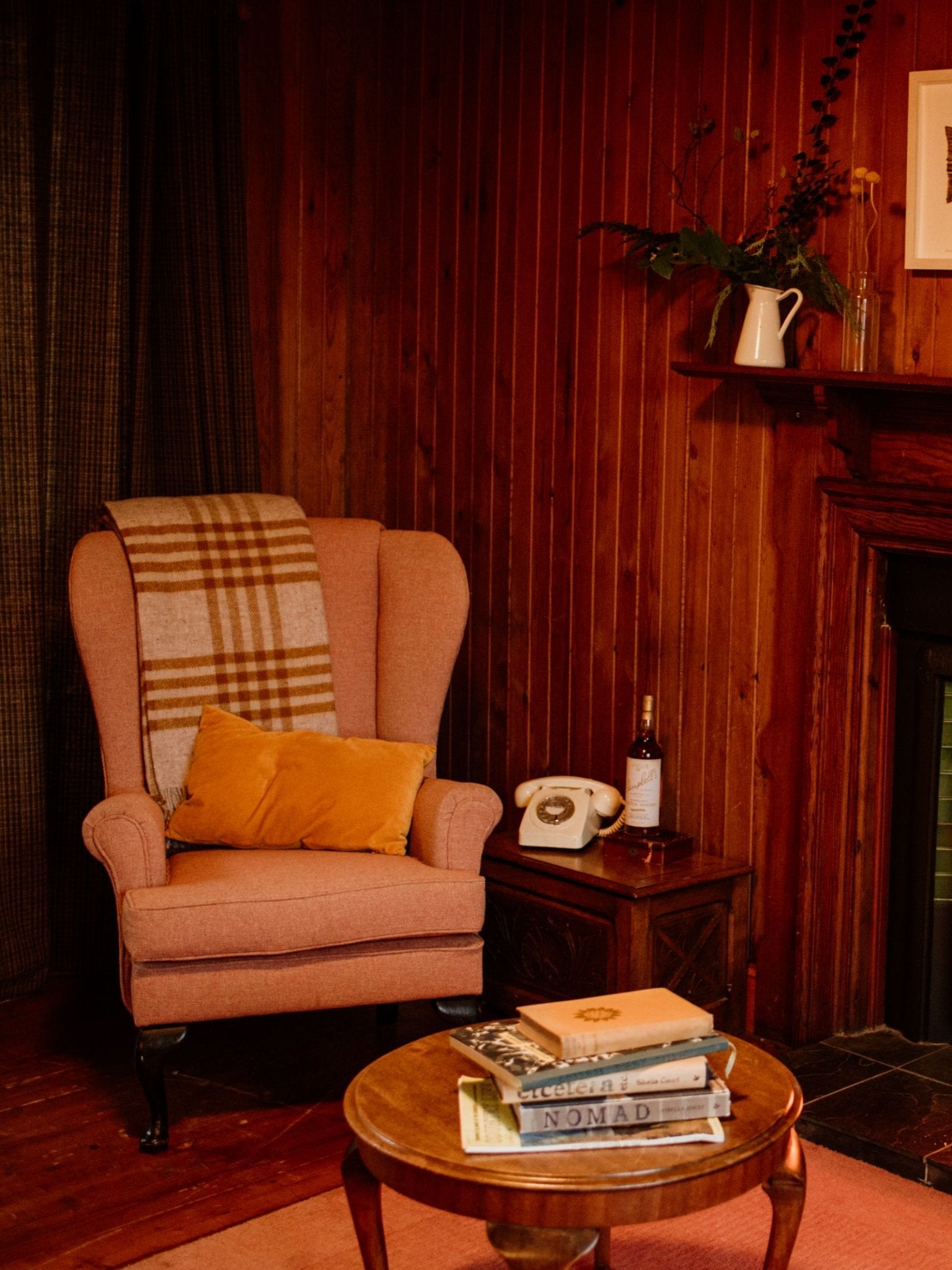 A cozy reading nook with a pink armchair, Campbell’s of Beauly Tarradale Lambswool Throw in classic check, mustard pillow, side table with vintage rotary phone, books, bottle, round coffee table, and wood-paneled walls reflecting timeless British style.