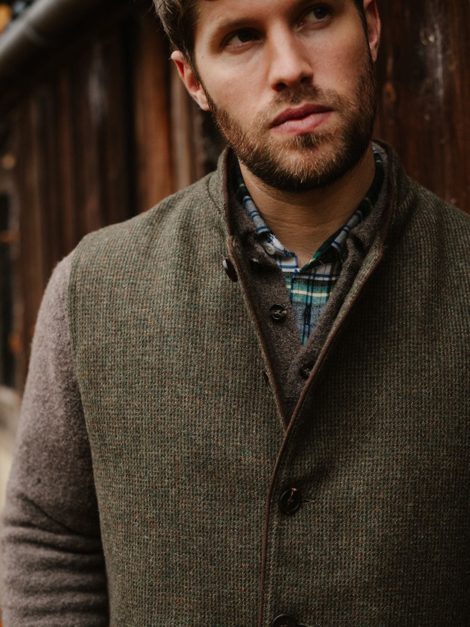 A man with a trimmed beard wears a Campbell's of Beauly Tweed Nehru Gilet over a plaid shirt, standing by a rustic wooden wall and glancing thoughtfully to the side.