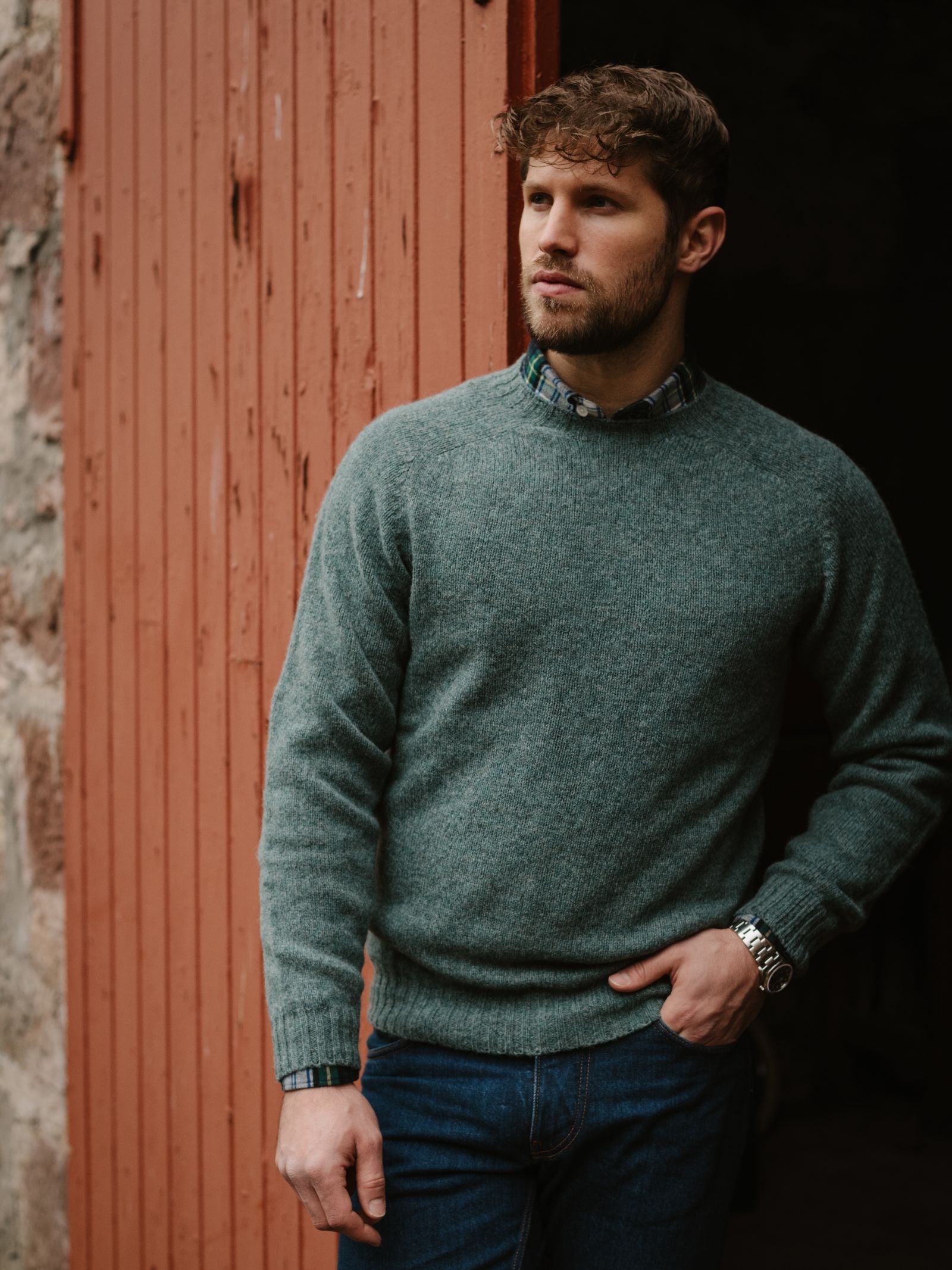 A man with curly brown hair and a beard stands before a red wooden door, wearing a green Campbell's of Beauly Shetland Jumper over a collared shirt and jeans, one hand in his pocket, looking to the side.