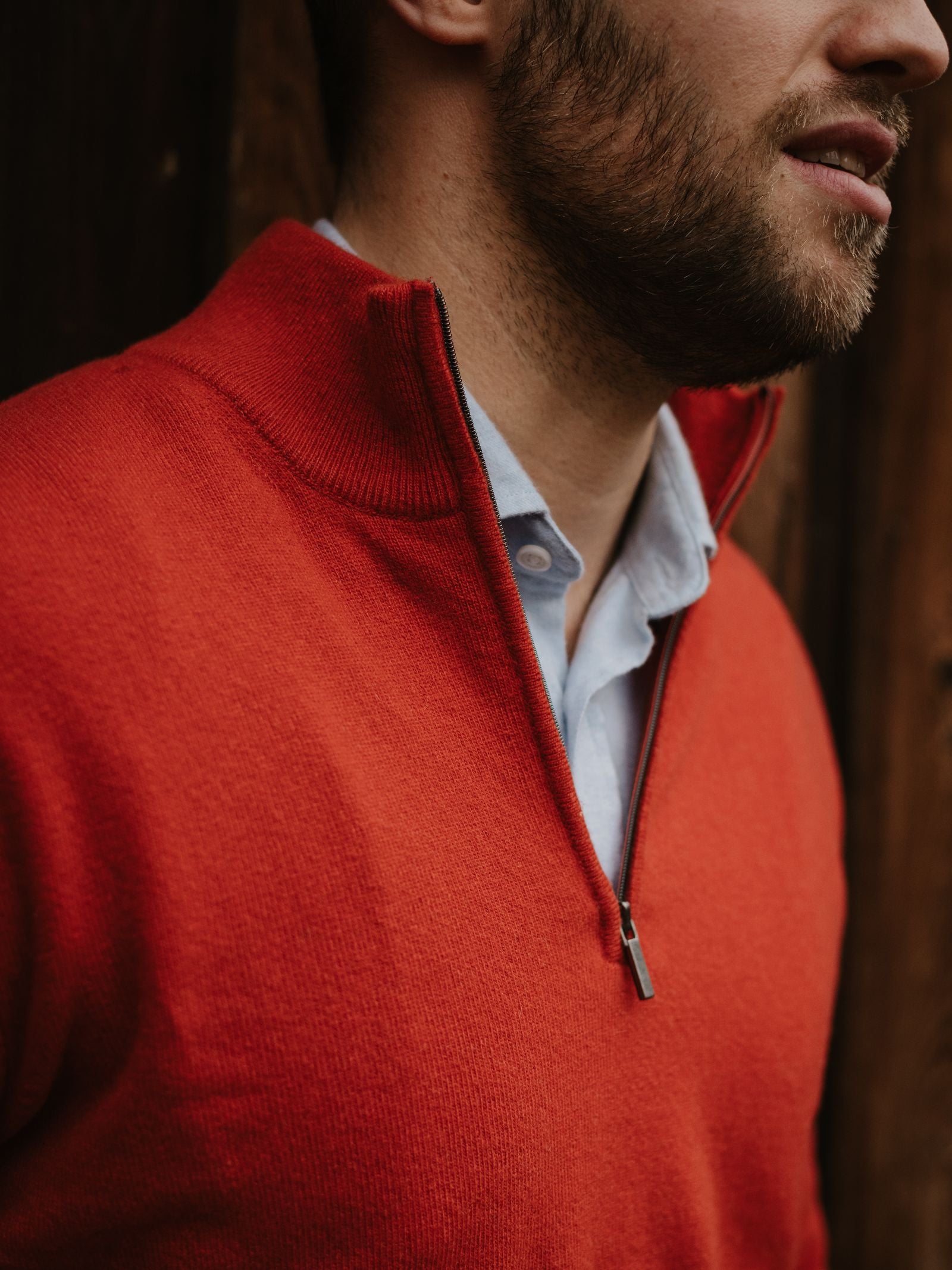 A man with a beard wears the Campbell's of Beauly Merino & Cashmere Quarter Zip Jumper in bright red over a light blue collared shirt, standing before a wooden background, his face partially visible as he looks to the side.