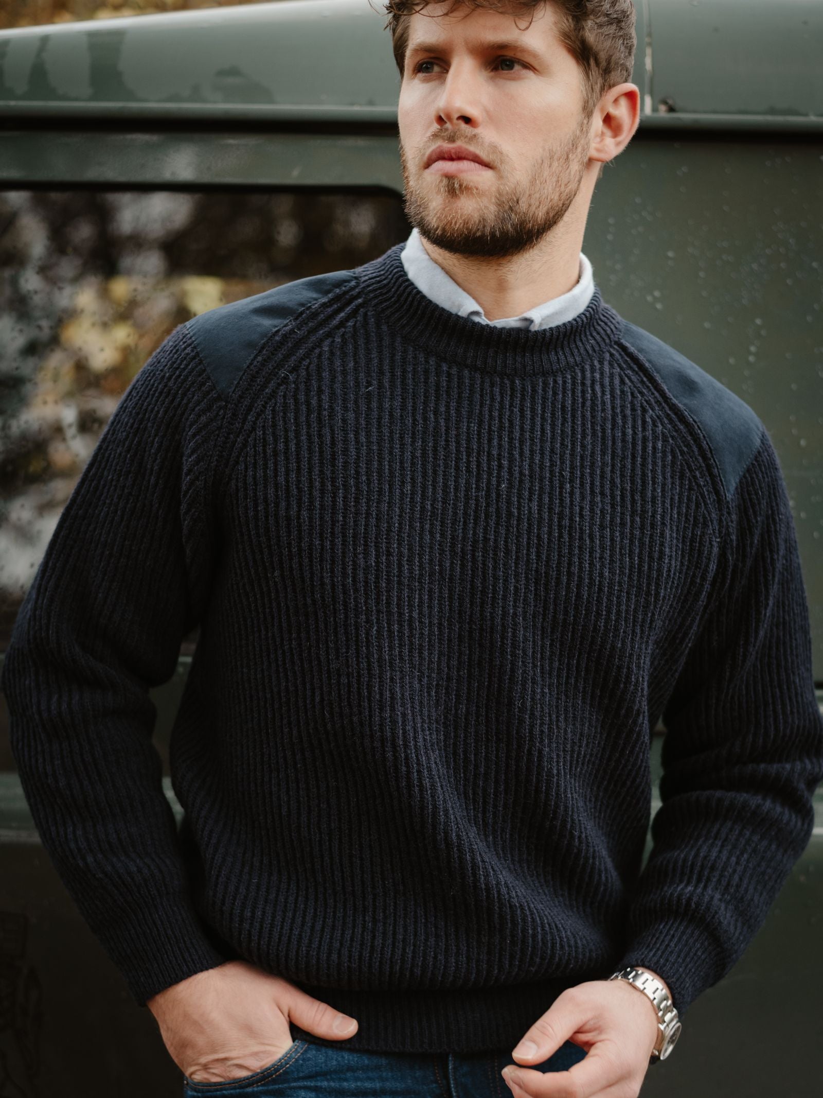 A man with short brown hair and a trimmed beard wears the Scotia Suede Jumper by Campbell's of Beauly over a collared shirt, standing outdoors beside a green vehicle with one hand in his pocket.