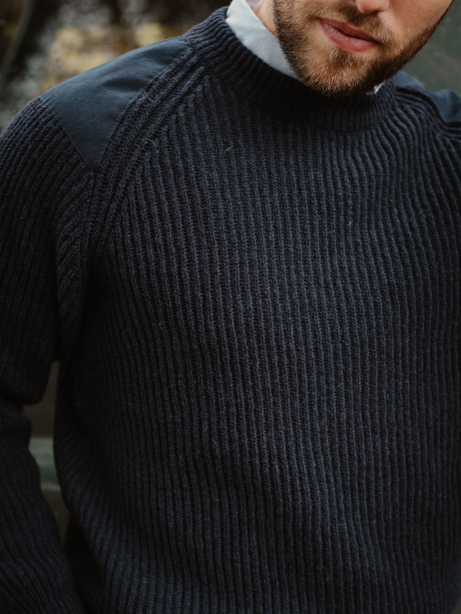 Close-up of a bearded person wearing the Campbell's of Beauly Scotia Suede Jumper—a thick, ribbed dark navy sweater with black shoulder patches—over a light collared shirt.