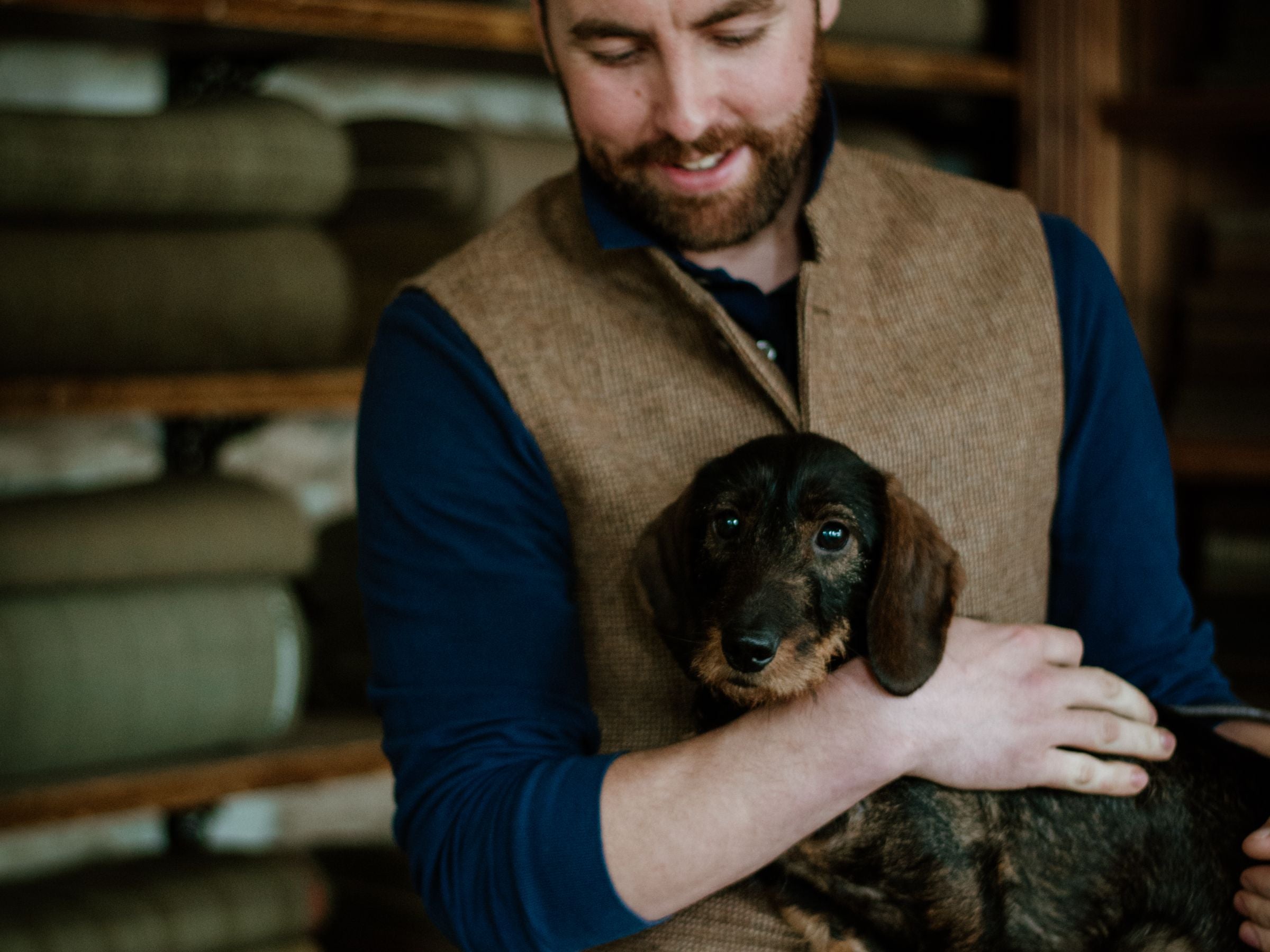 A man with a brown vest and navy shirt gently holds a brown dachshund puppy in his arms, smiling warmly. Shelves with stacked fabric rolls are blurred in the background.