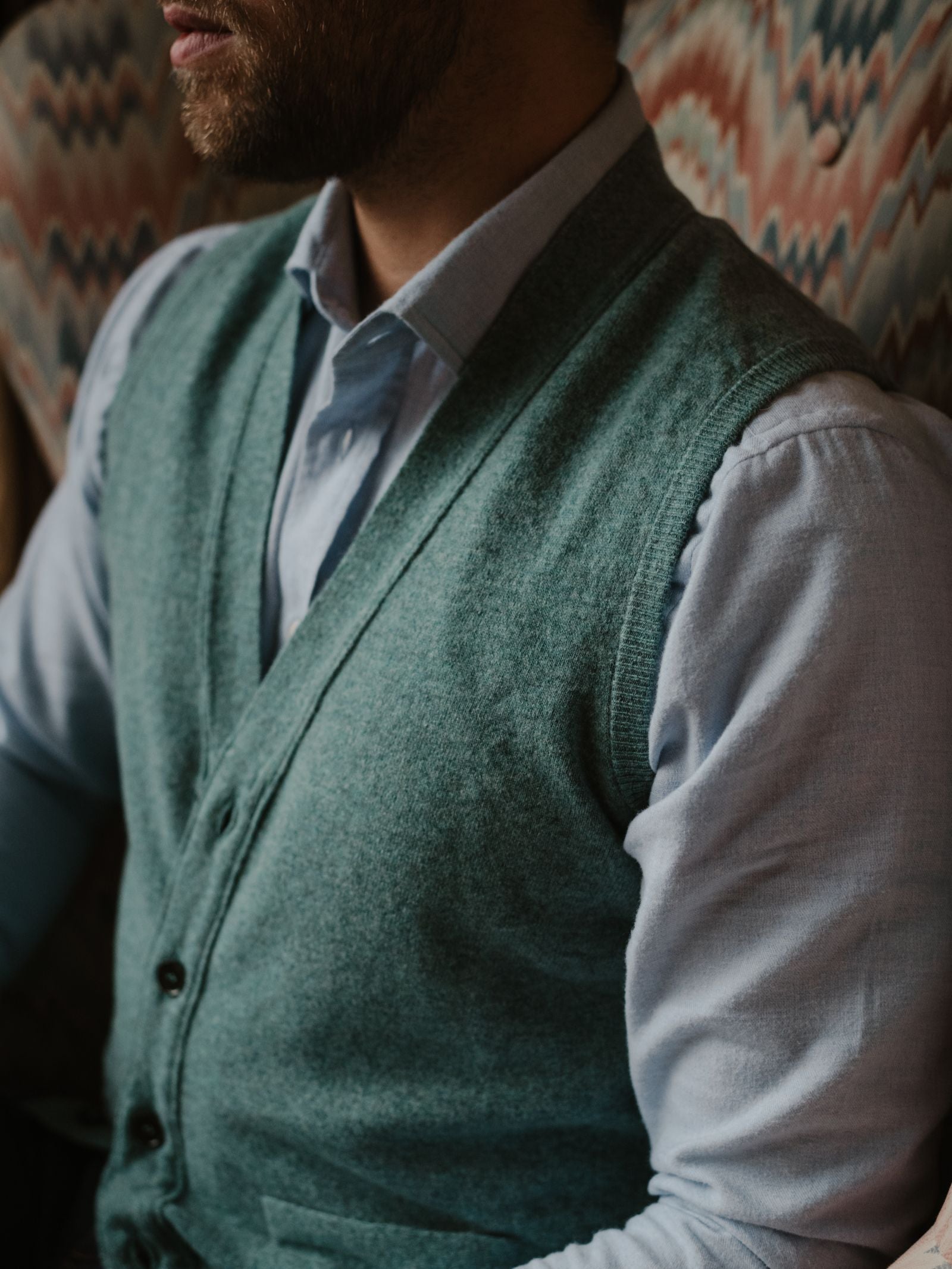 A man wearing a light blue dress shirt and the Geelong Button Waistcoat by Campbell's of Beauly sits on a patterned chair, his upper body and part of his face visible in the cropped image.