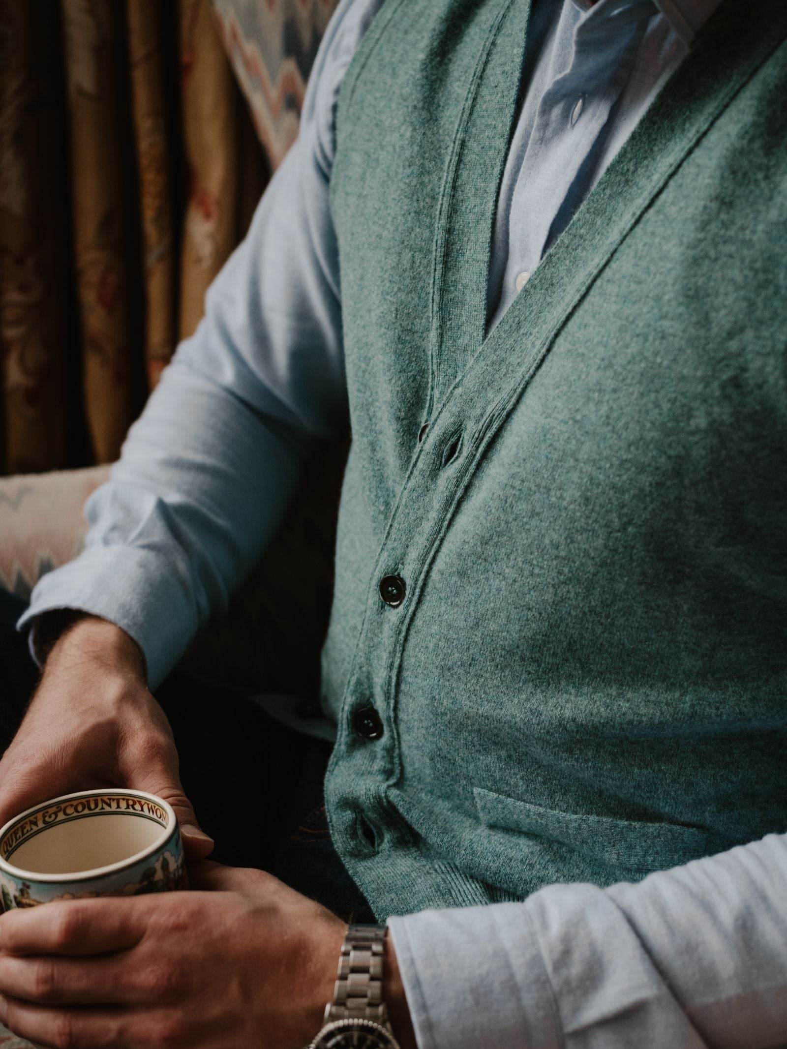 A person in a light blue shirt and a Campbell's of Beauly Geelong Button Waistcoat sits holding a patterned mug in their left hand. Their face is hidden, and they wear a silver wristwatch.