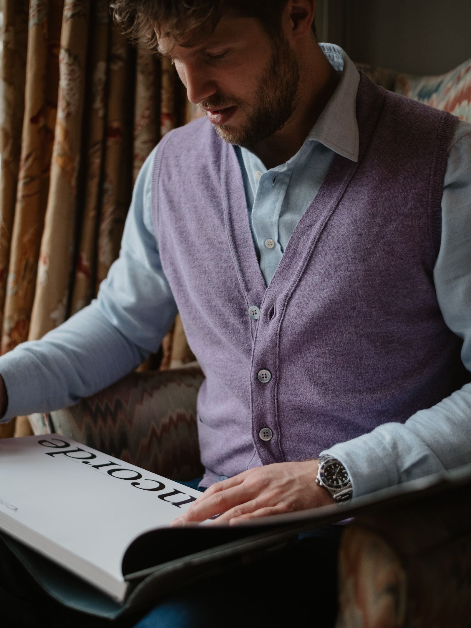 A man in a light blue shirt and Campbell's of Beauly Auld Stock Geelong Button Waistcoat sits in an armchair, reading a large magazine near patterned curtains—adding elegance to his country wardrobe.