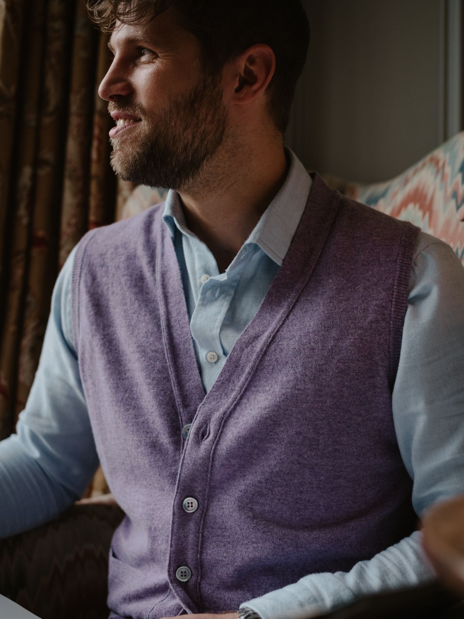 A bearded man in a light blue shirt and Campbell’s of Beauly Auld Stock Geelong Button Waistcoat sits indoors, smiling to the side. Patterned curtains and a zigzag chair enhance his charming country wardrobe scene.